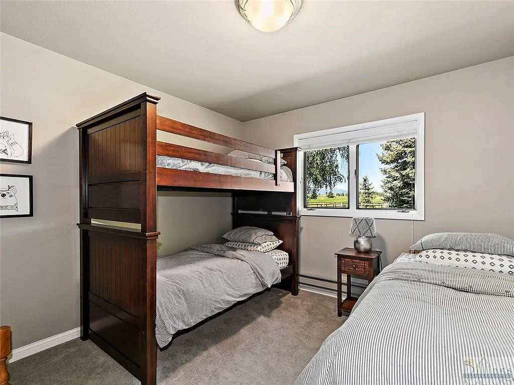 Bedroom with a wooden bunk bed, a single bed, a nightstand with a lamp, and a window showing trees and a yard outside.