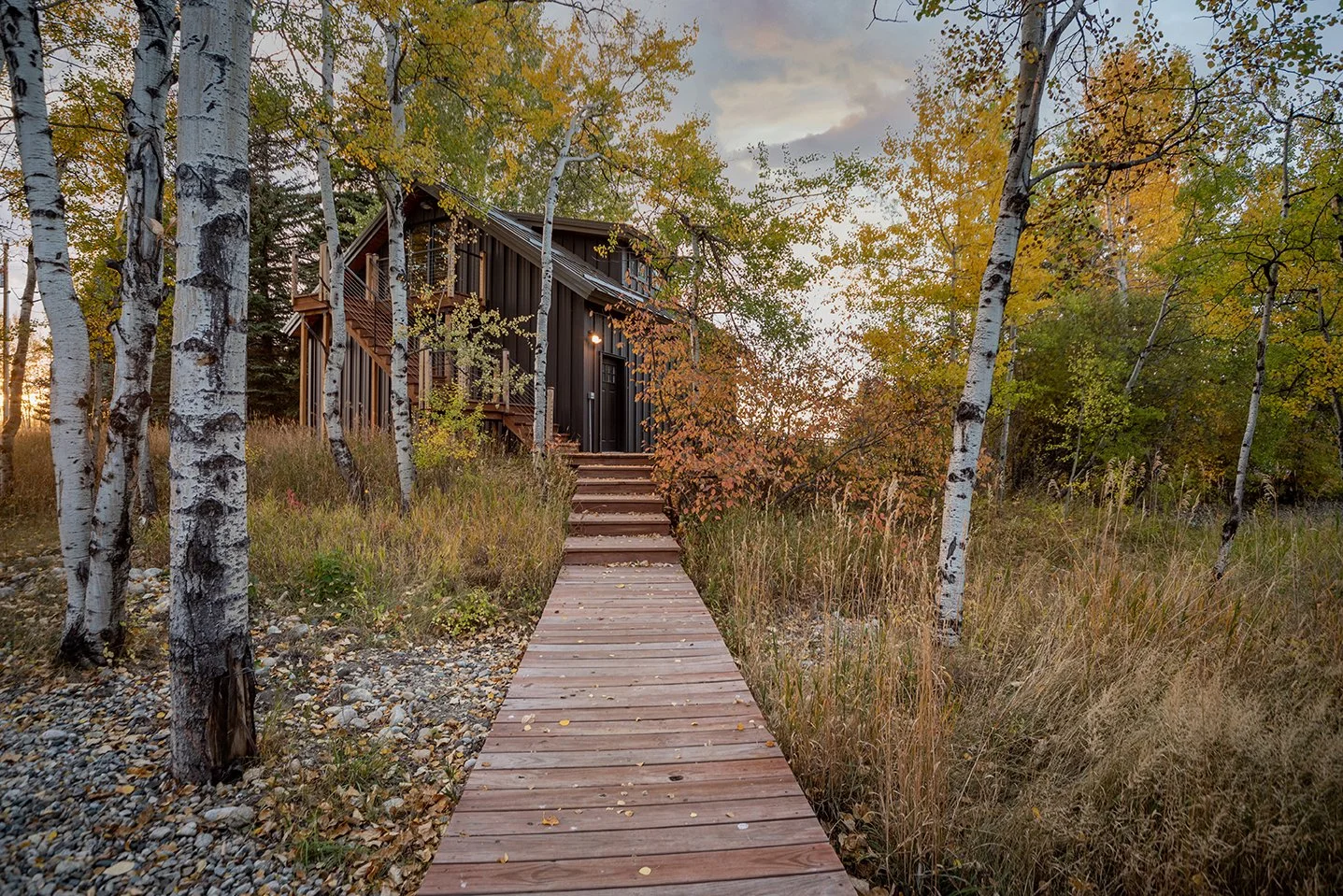 A wooden walkway leading to a dark-colored house surrounded by trees with autumn leaves.