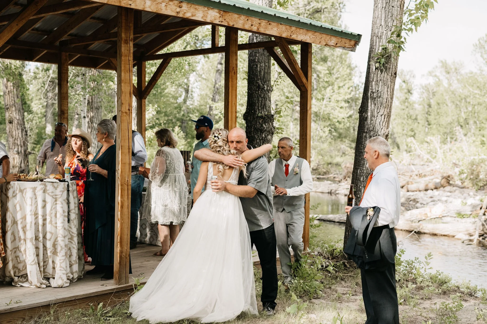 A bride and groom hug on a wooden outdoor wedding porch surrounded by guests in a wooded area near a stream.