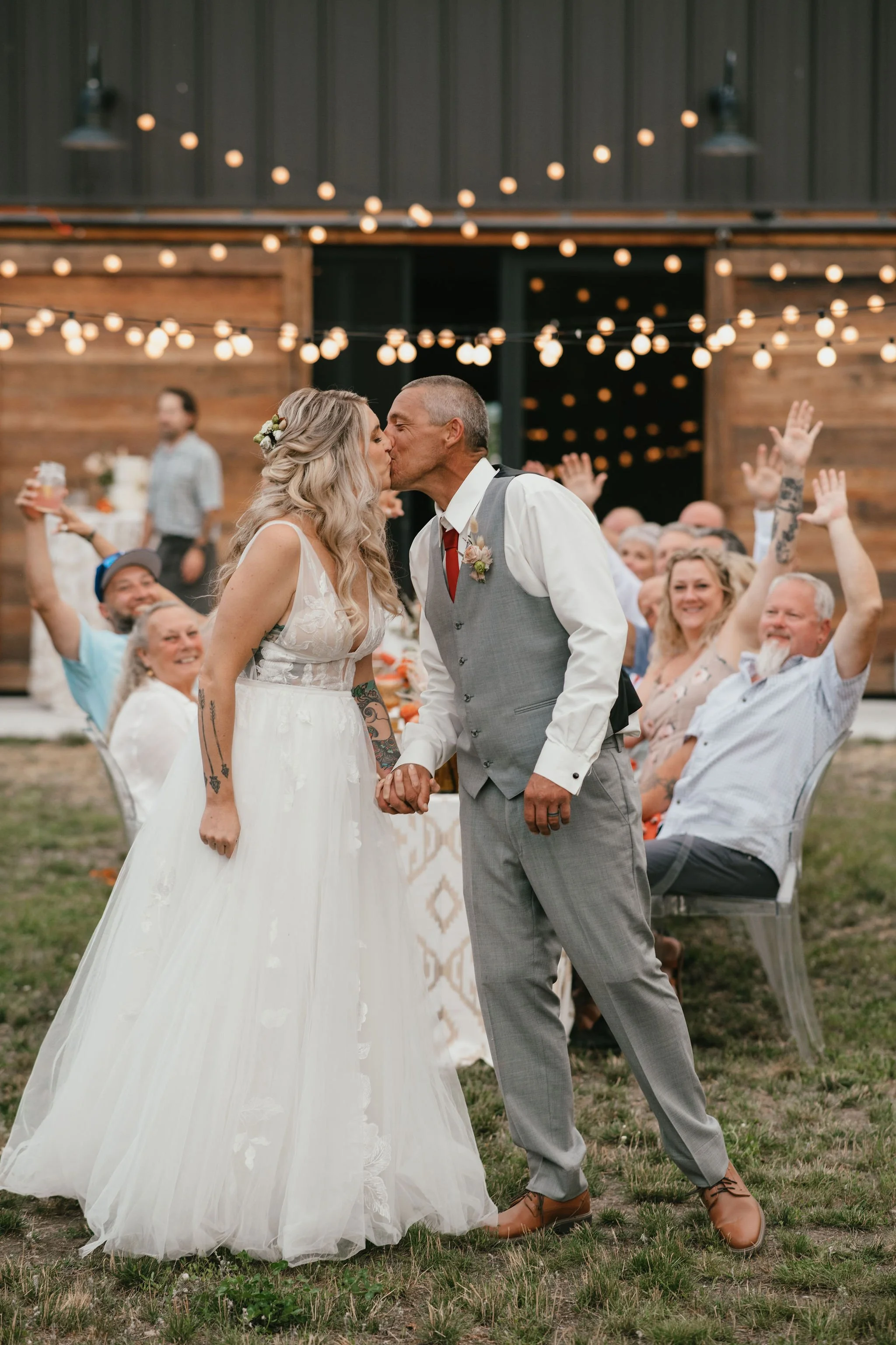 Couple in wedding attire kissing, holding hands at outdoor wedding reception with guests seated at tables in background