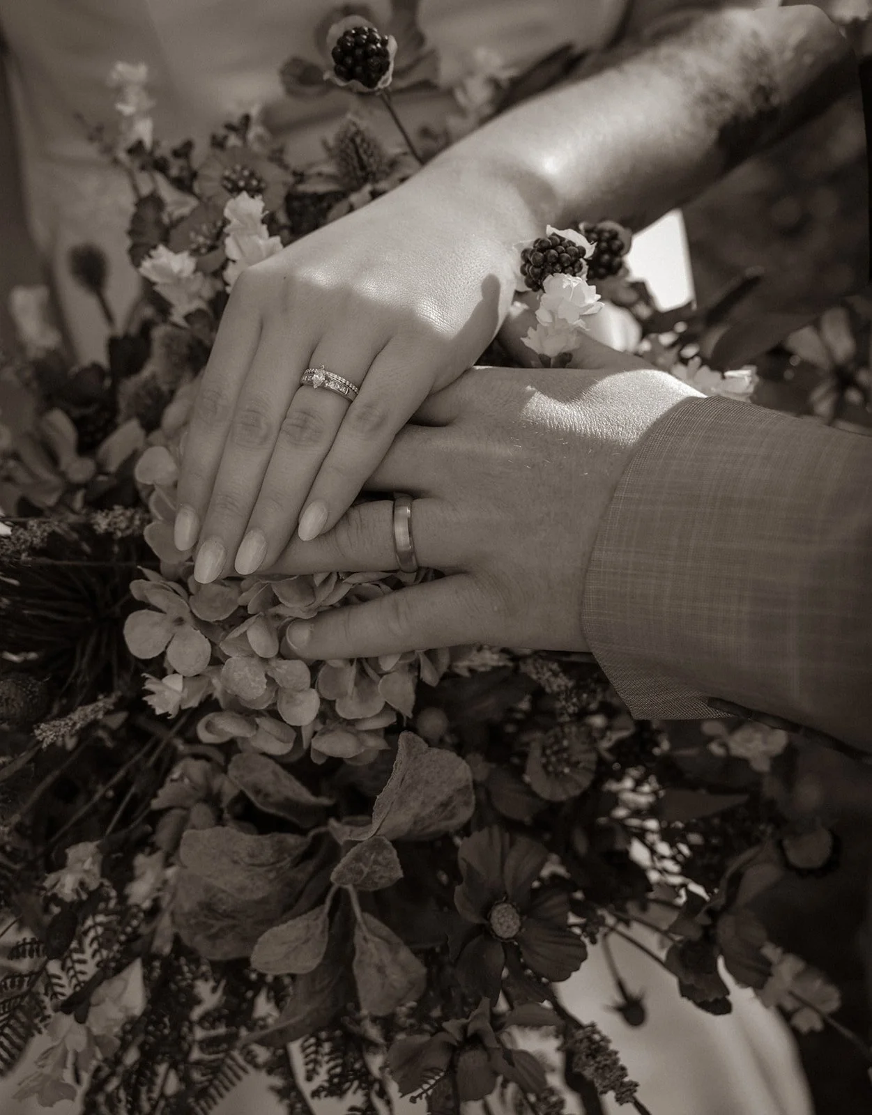Close-up of a couple's hands with wedding rings, resting on a bouquet of flowers, black and white photograph.