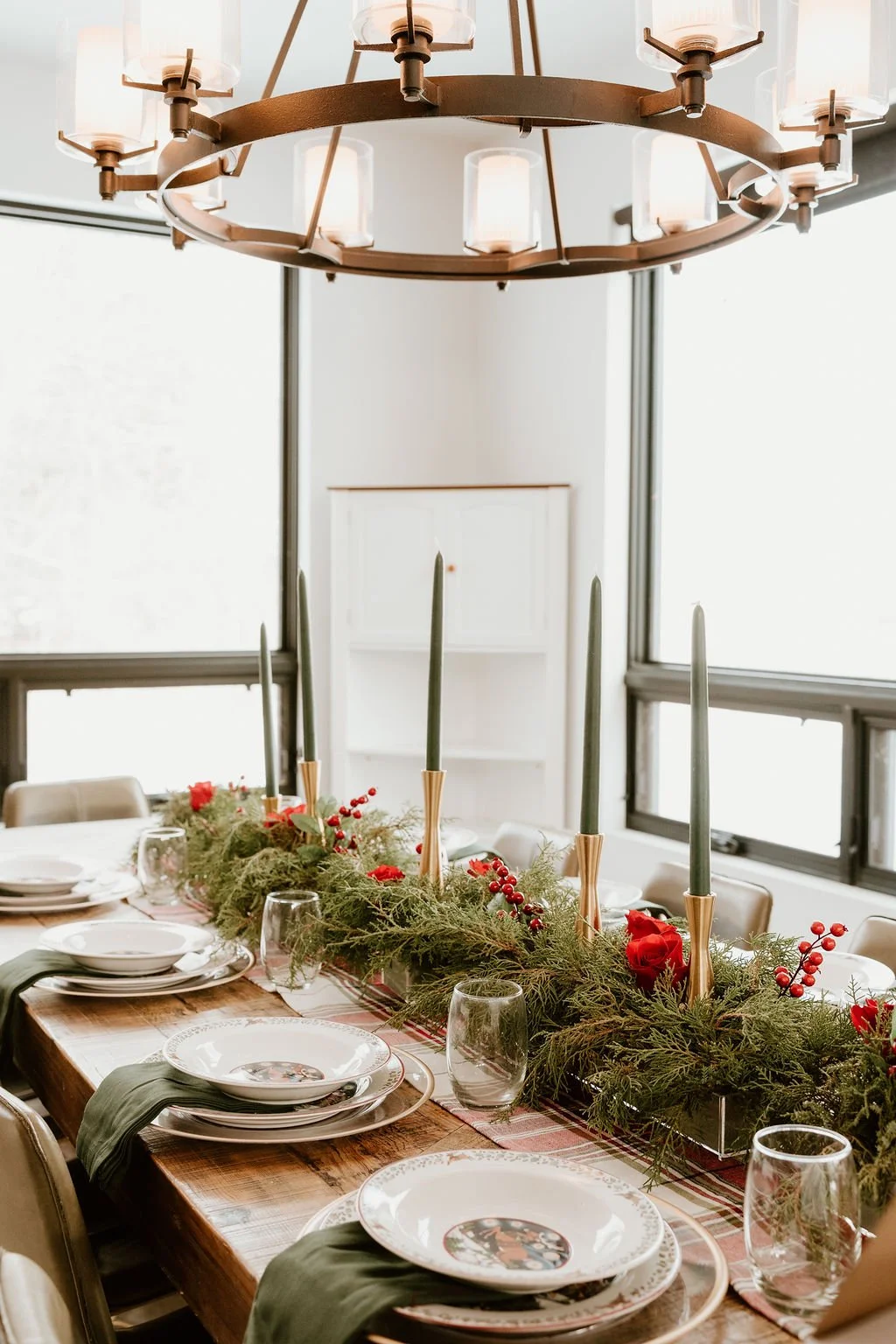 A dining table decorated for a holiday with a greenery garland, red berries, candles, and plates and glasses.