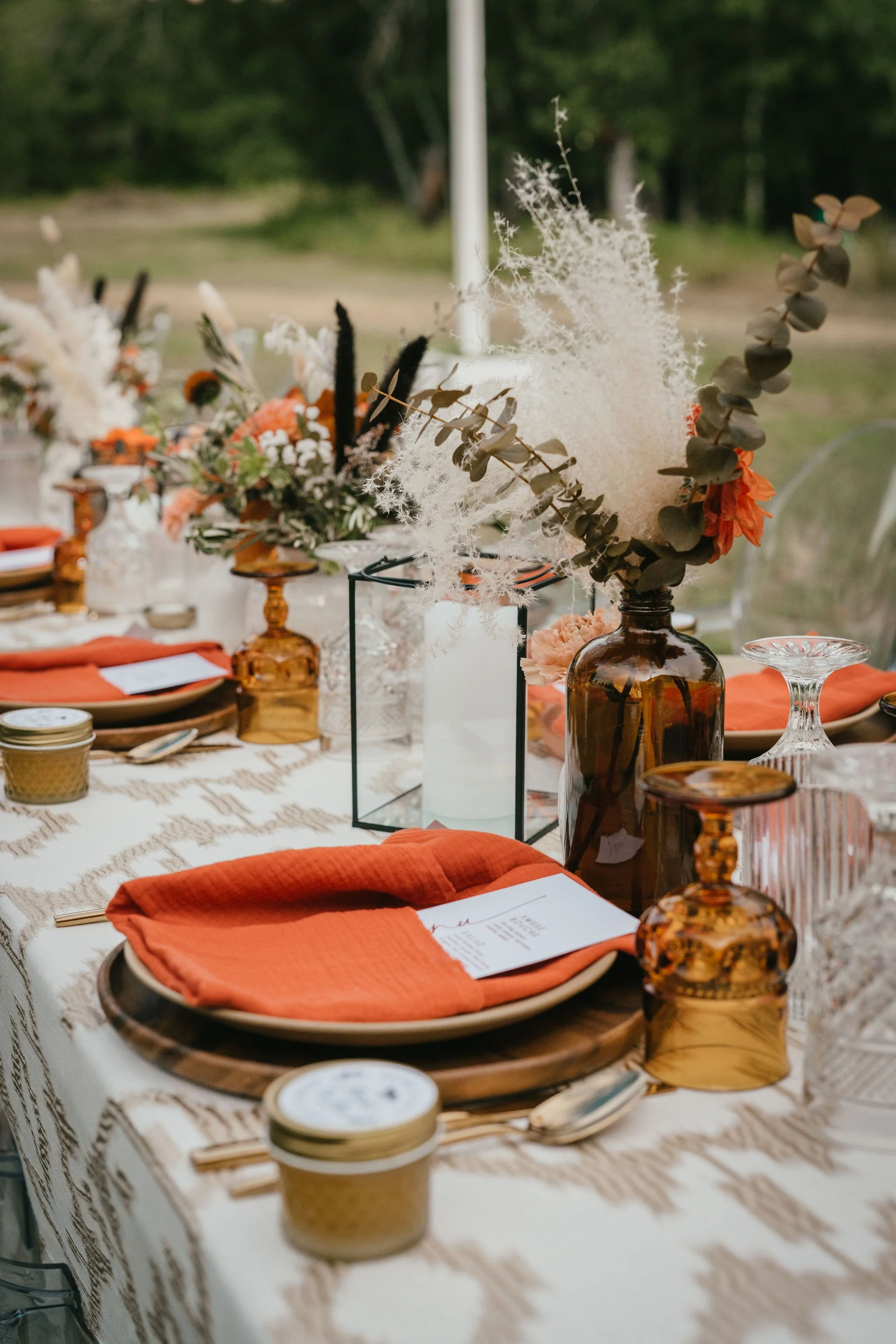 Elegant outdoor table setting with amber glass vases, white candles, and peach-colored napkins on beige patterned tablecloth, decorated with floral arrangements including dried foliage and pampas grass.
