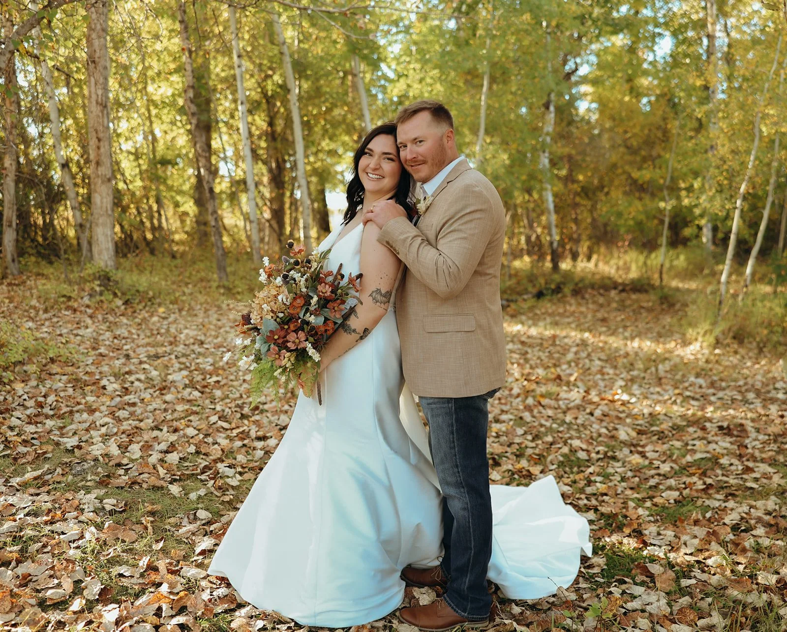 A bride and groom standing together outdoors in a wooded area during autumn. The bride is holding a large bouquet and wearing a white wedding dress, while the groom is in a beige blazer and jeans. They are smiling and embracing.