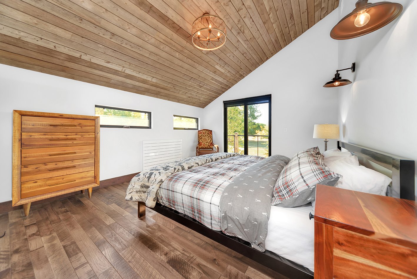A bedroom with a wooden vaulted ceiling, hardwood flooring, a bed with plaid and patterned bedding, a wooden nightstand, a lamp, a cushioned chair, and a sliding glass door leading to an outdoor balcony.