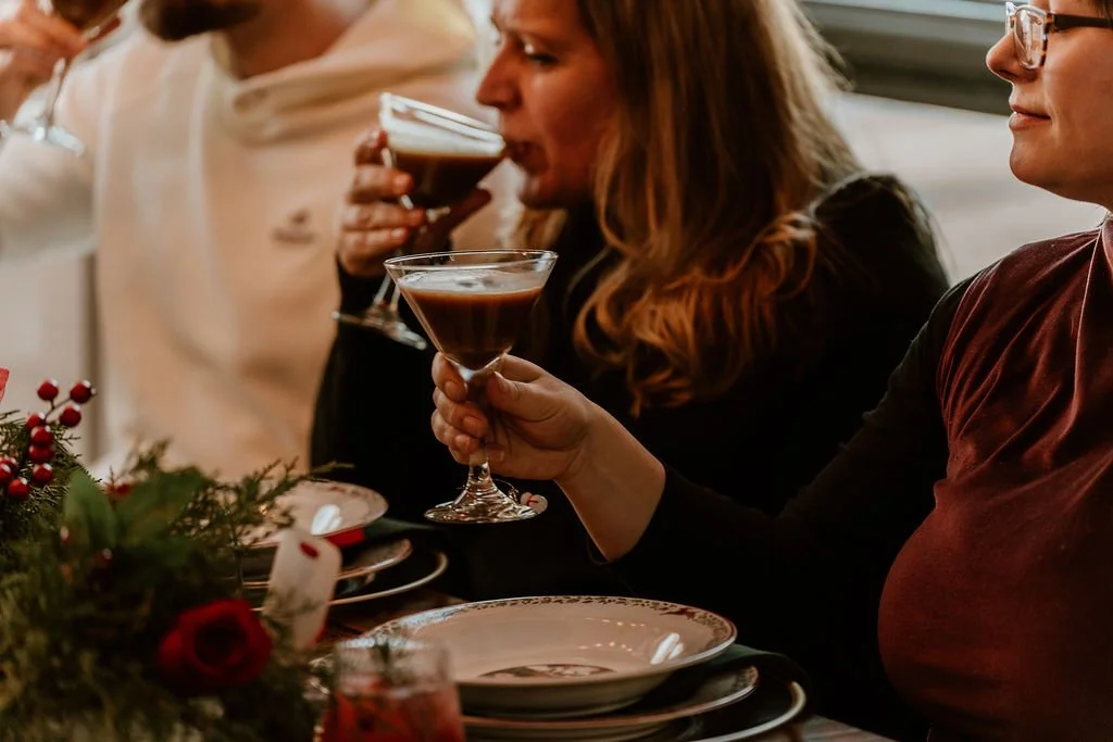 People sitting at a festive dining table, holding and drinking cocktails, with holiday decorations and plates in the foreground.