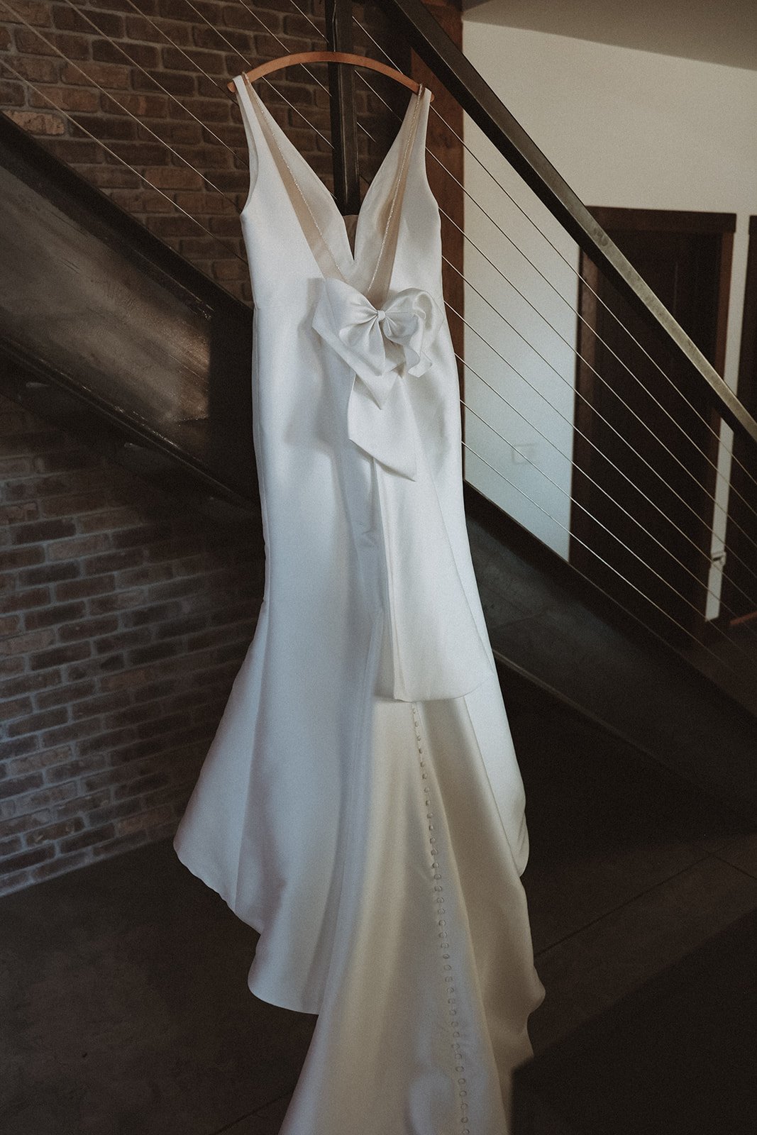 White wedding dress with a large bow hanging on a hanger near a staircase with a brick wall in the background.
