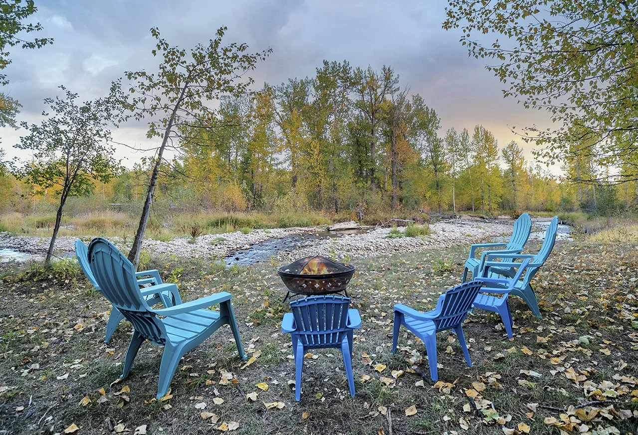 Six blue Adirondack chairs arranged in a circle around a fire pit on a grassy area near a river, with trees and autumn leaves in the background.