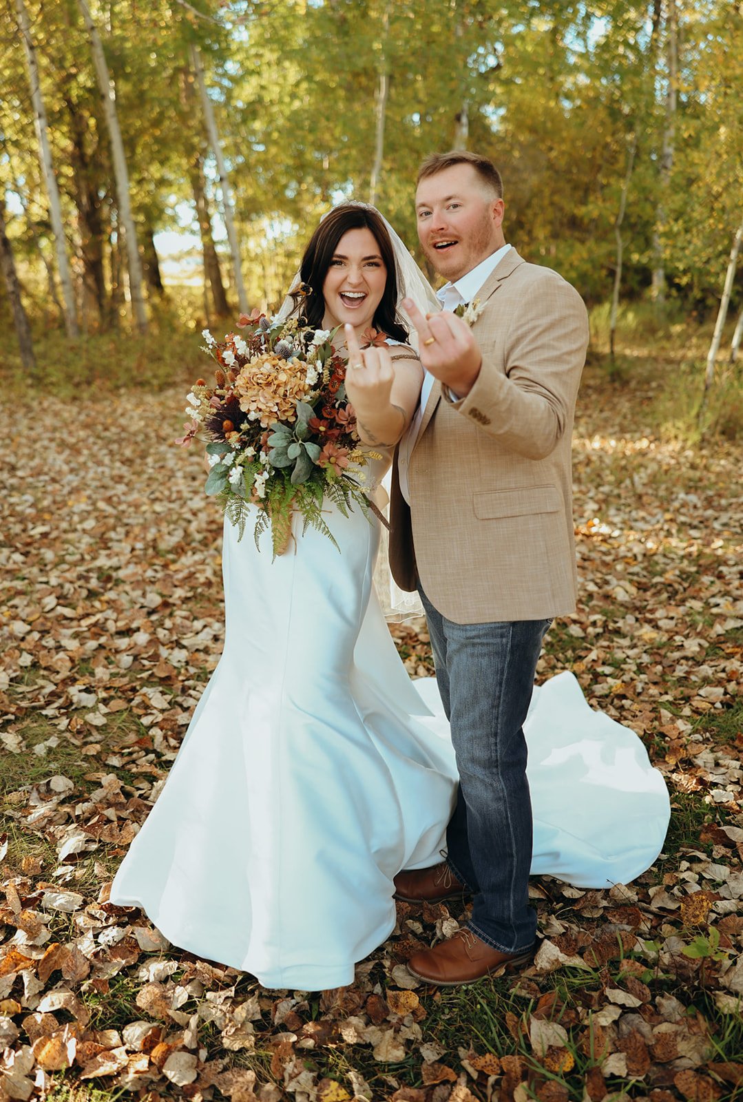 A smiling bride in a white wedding dress holding a bouquet, and a groom in a tan blazer and jeans, standing together outdoors among fallen autumn leaves and trees, making playful gestures.