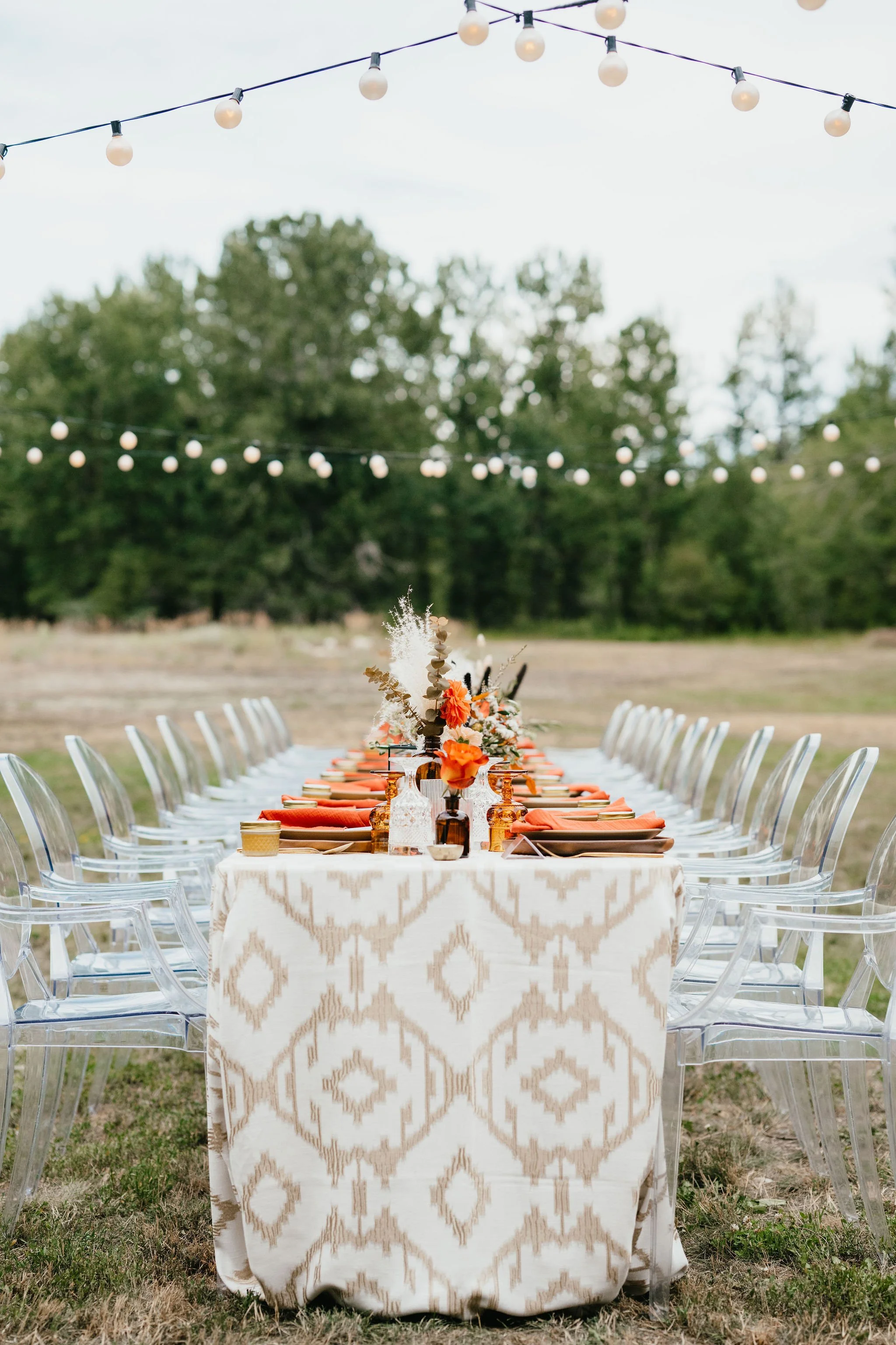 Outdoor table setting for a celebration with floral centerpieces and string lights overhead.