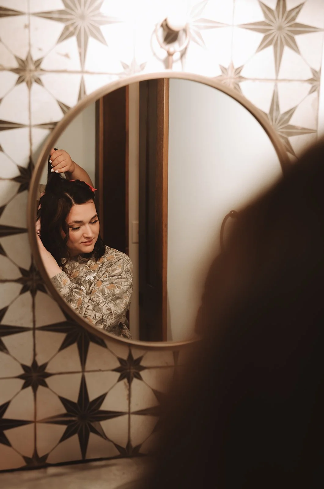 A woman with dark hair in curlers looking at herself in a small round mirror, adjusting her hair in a room with decorative tiled walls.