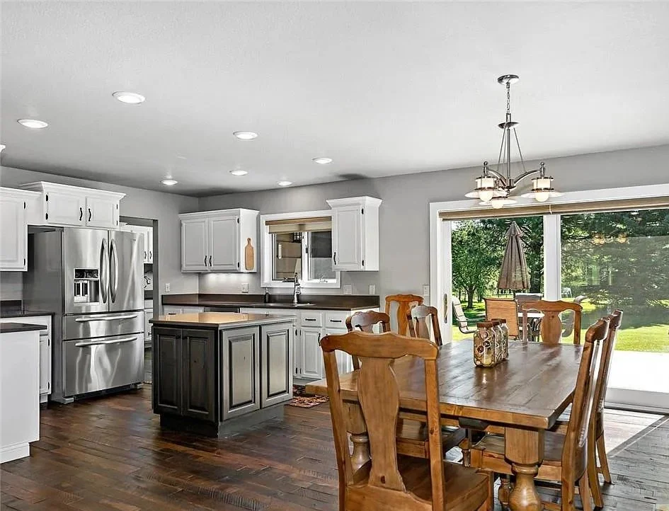 Kitchen with white cabinets, stainless steel refrigerator, black countertops, and a center island. Wooden dining table with six chairs and a chandelier. Large window and sliding glass door leading to a backyard with trees and grass.
