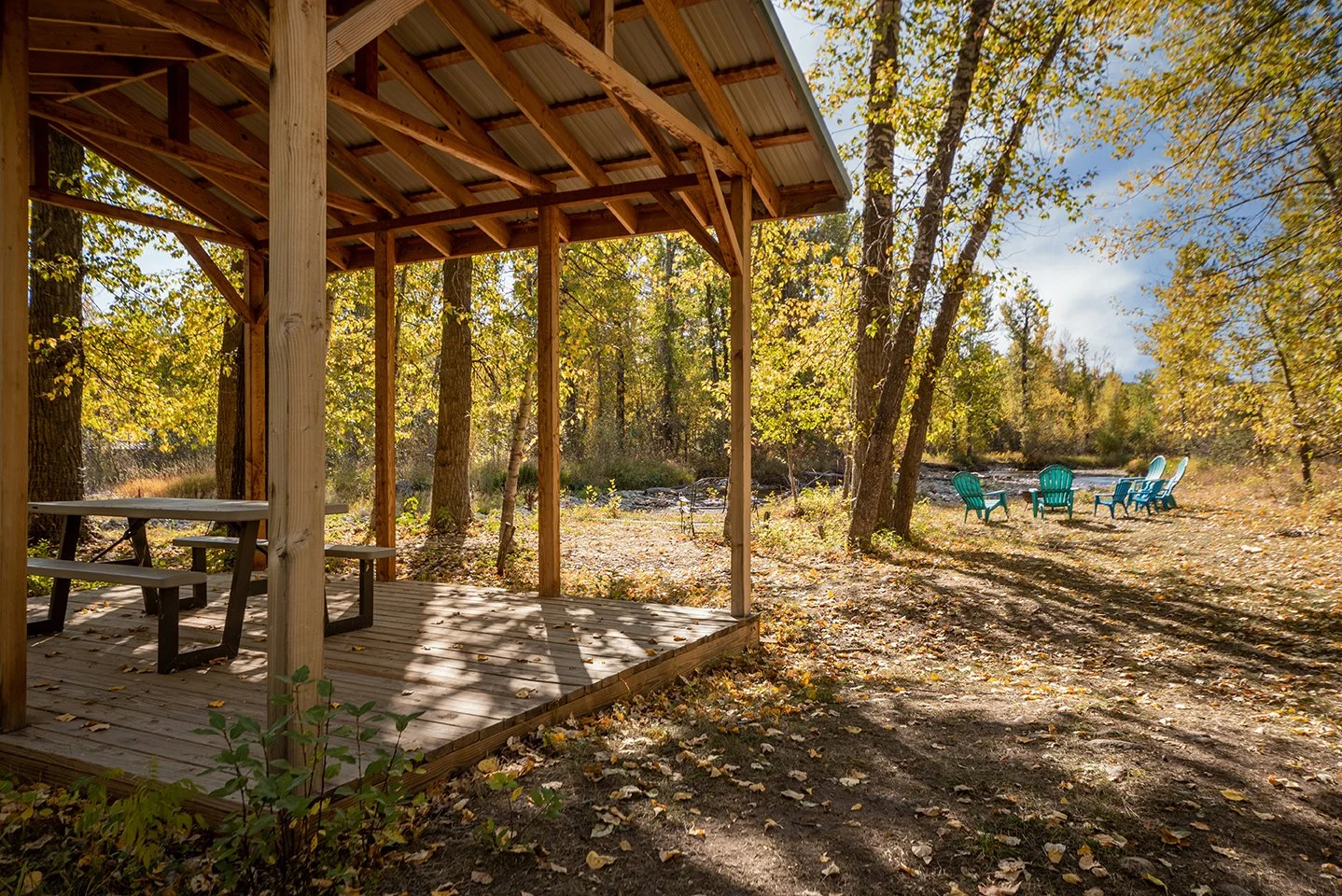 A wooden pavilion with a picnic table is in a forested area with many trees and fallen leaves. In the background, there are several colorful chairs arranged on the ground. The scene is illuminated by sunlight filtering through the trees.