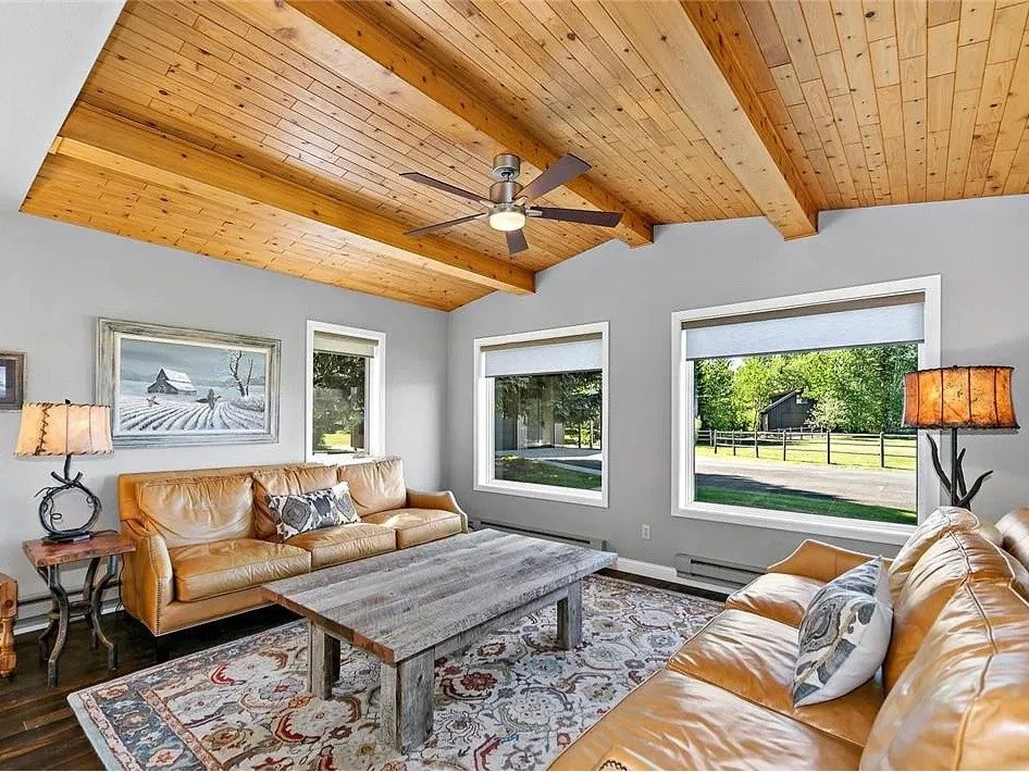 Living room with light gray walls, wooden ceiling with beams, ceiling fan, two large windows with white blinds, leather sofas, a wooden coffee table, area rug, side table with a lamp, and a painting of a winter landscape on the wall.