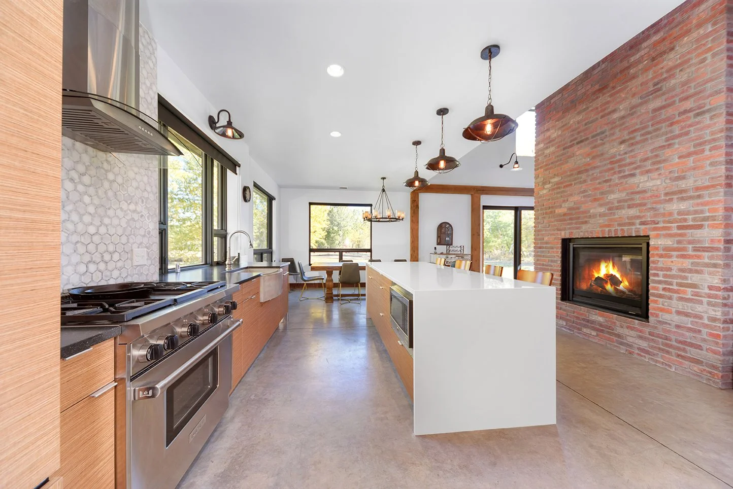 Open-concept kitchen with a brick fireplace, white island, stainless steel range, wood cabinets, black windows, and modern pendant lighting, with a dining area in the background.