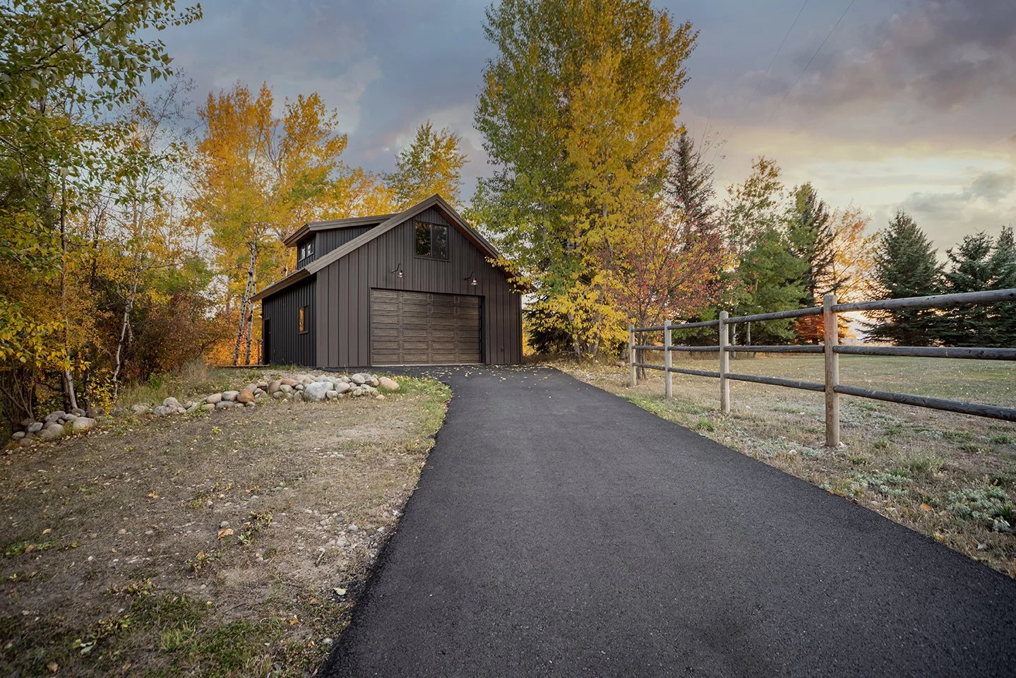 A dark gray barn with an attached garage on a paved driveway, surrounded by autumn trees with colorful leaves, a wooden fence to the right, and a sky with clouds.