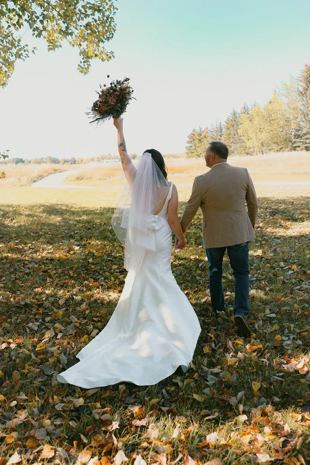 A bride and groom walking hand in hand outdoors during autumn, with the bride raising a bouquet of flowers in her right hand, wearing a white wedding gown and veil, in a scenic park with fallen leaves and trees.