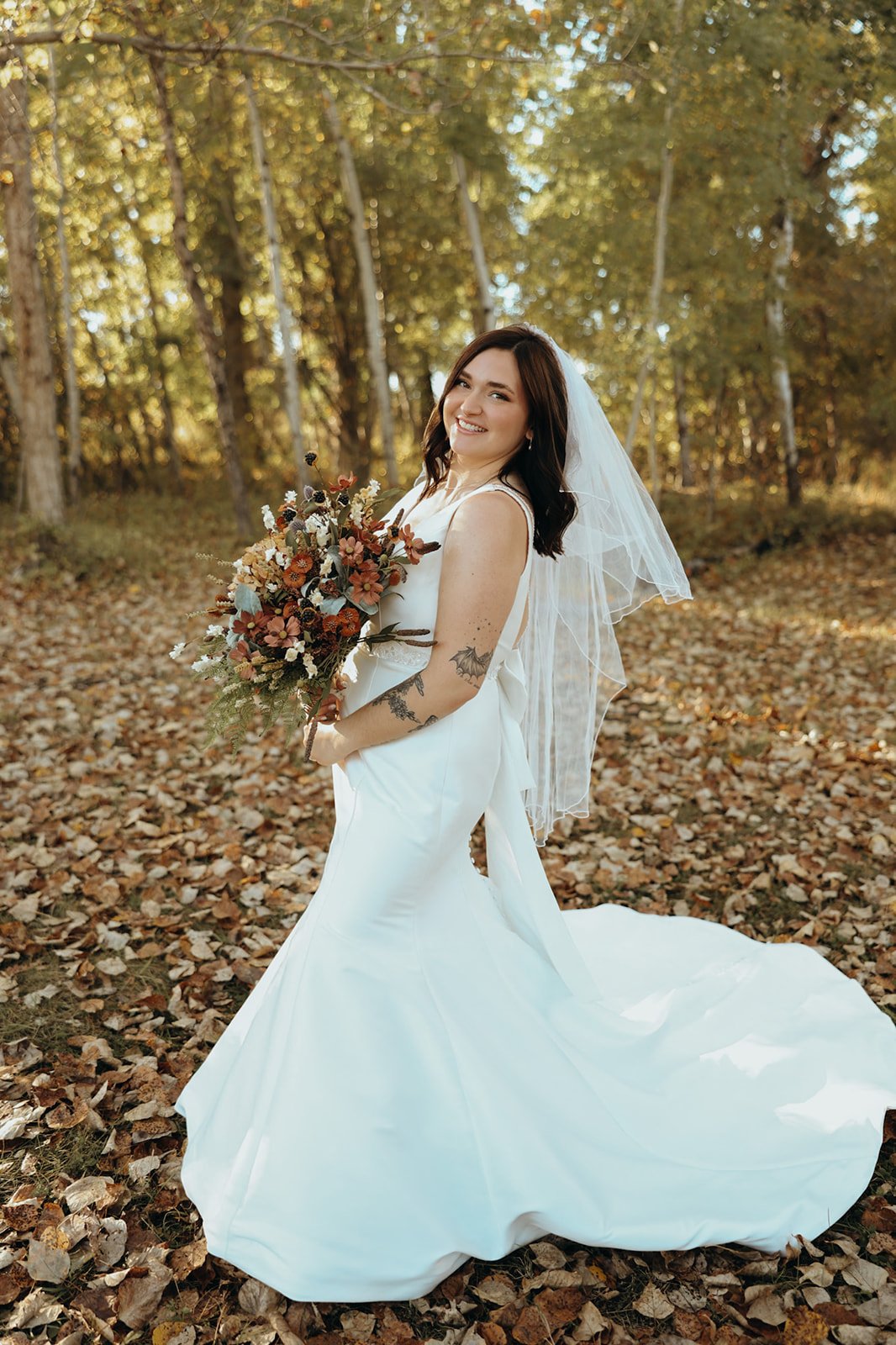 A woman in a white wedding dress with a veil, holding a colorful bouquet of flowers, standing outdoors among fallen autumn leaves in a wooded area, smiling at the camera.