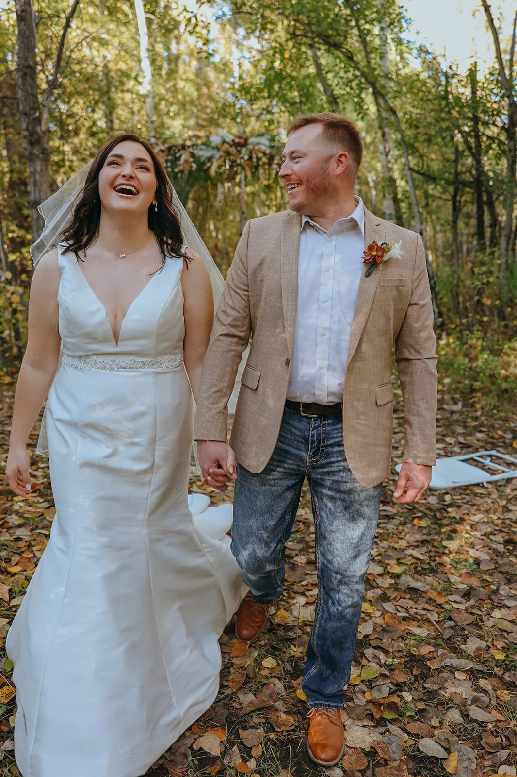 A bride and groom walking hand-in-hand outdoors among trees with fallen leaves, celebrating their wedding day.