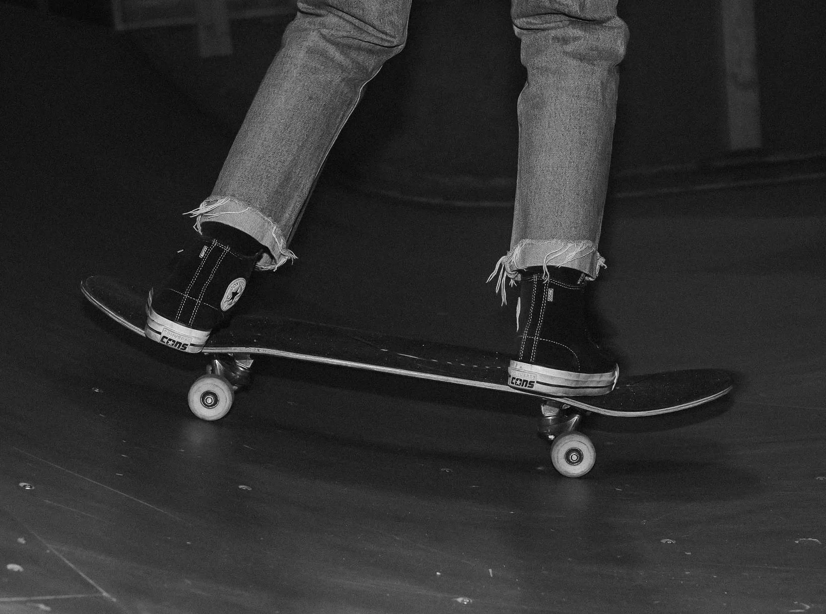 A person standing on a skateboard, wearing black high-top Converse sneakers and distressed jeans, on a dark surface.