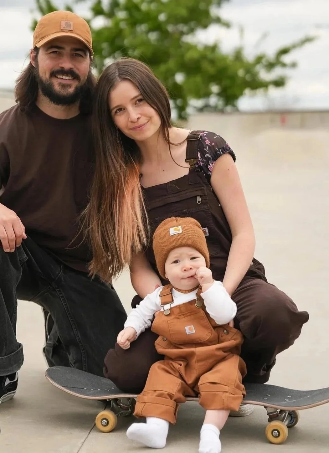 A family of three outdoors on a cloudy day, with a man, a woman, and a baby in brown Carhartt clothing and hats, sitting on a skateboard, with a green tree in the background.