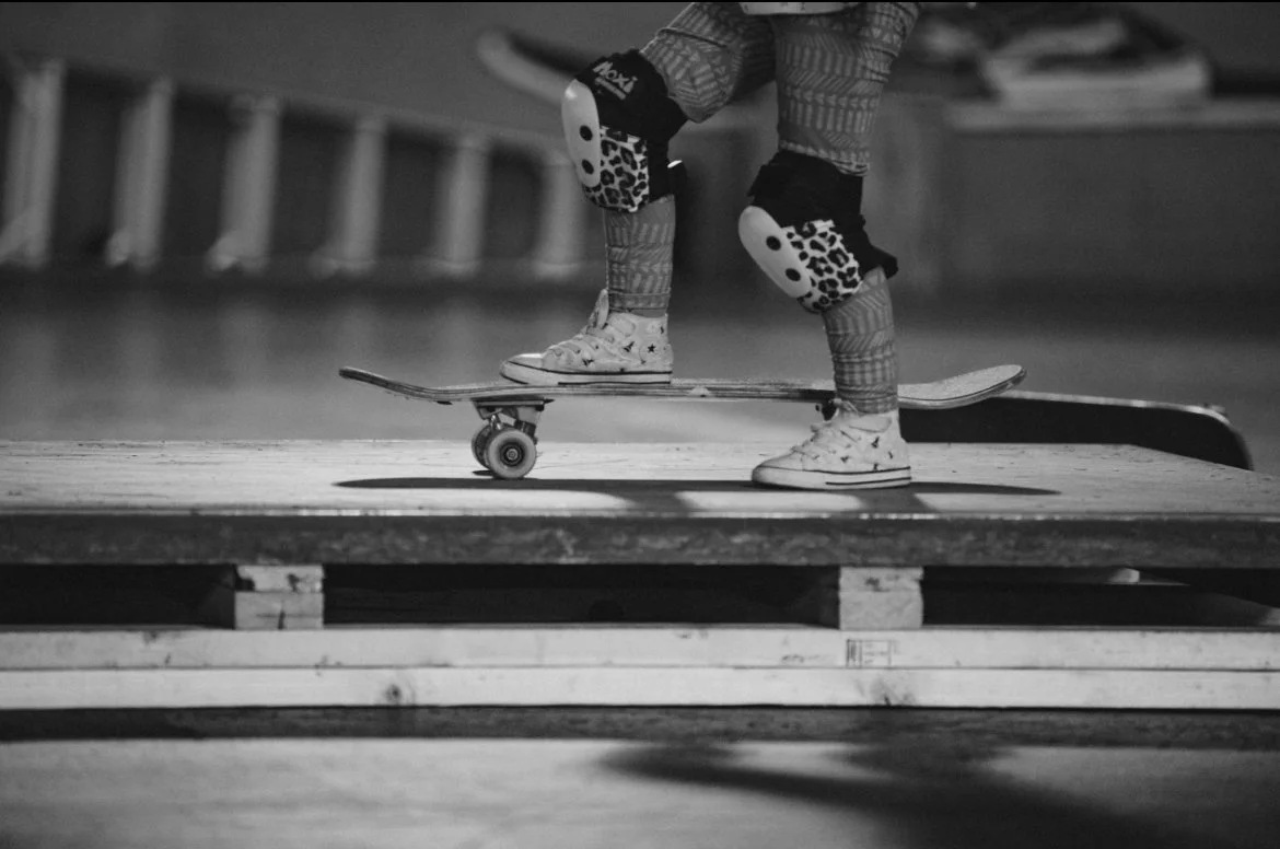 Close-up of a skateboarder’s legs in protective gear standing on a skateboard at a skatepark, with a ramp below.