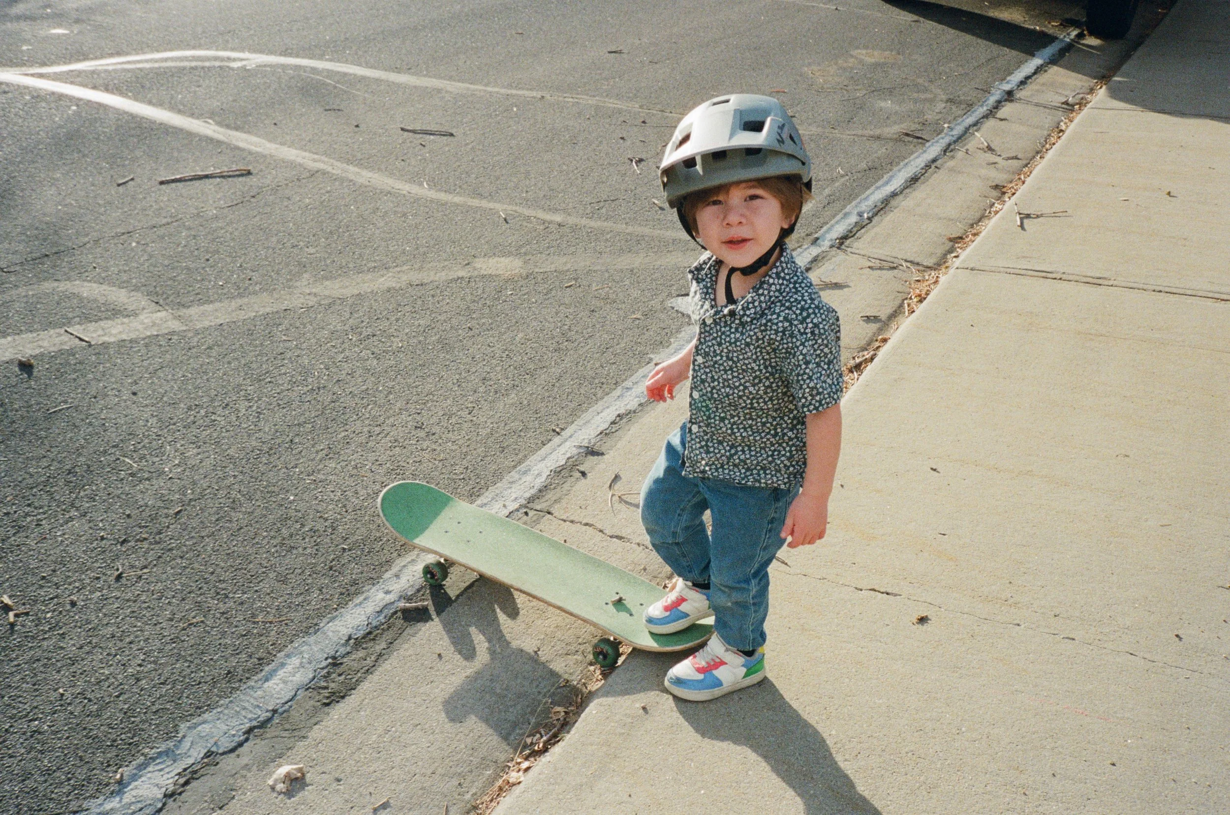 Young boy wearing a helmet standing on sidewalk next to skateboard.