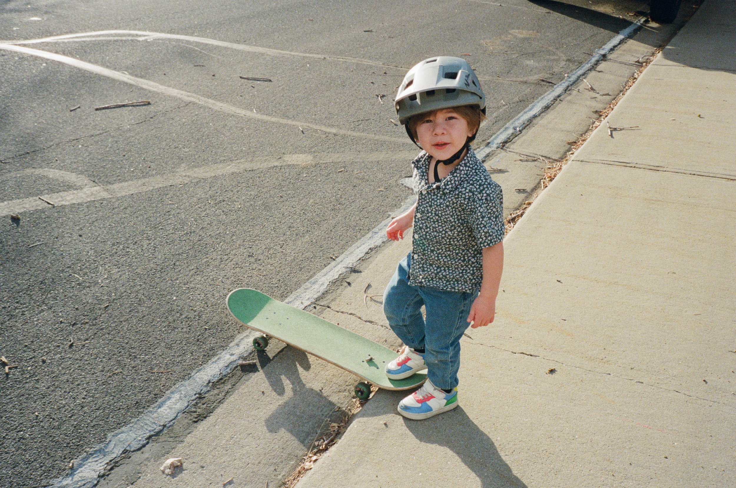 A 3 year old turns to look at the camera as he puts one foot on the skateboard, ready to ride