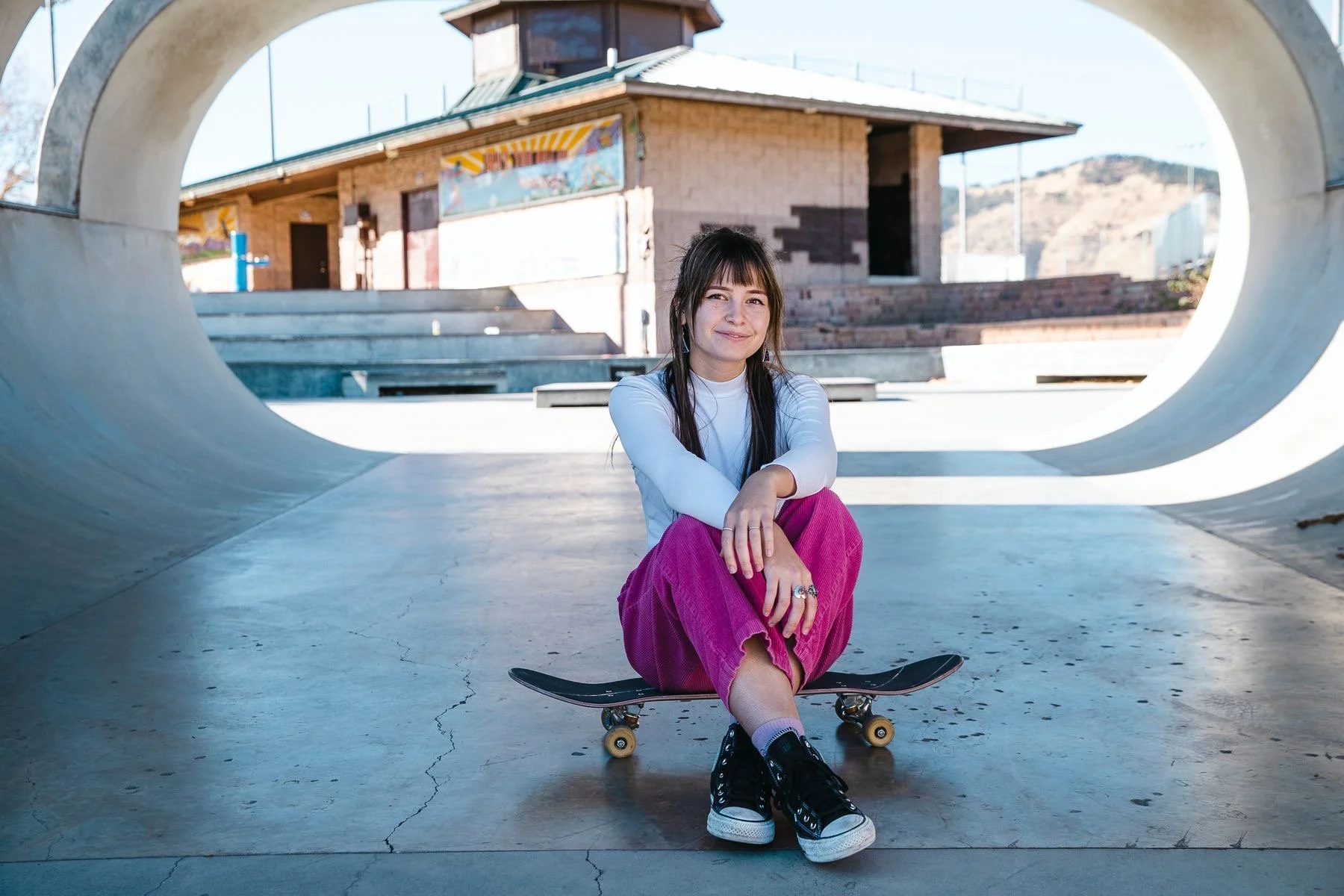 A young woman with dark hair, wearing a white long-sleeve shirt and pink corduroy pants, sits on a skateboard underneath a concrete skatepark sculpture shaped like a tunnel, with a building and hills in the background.