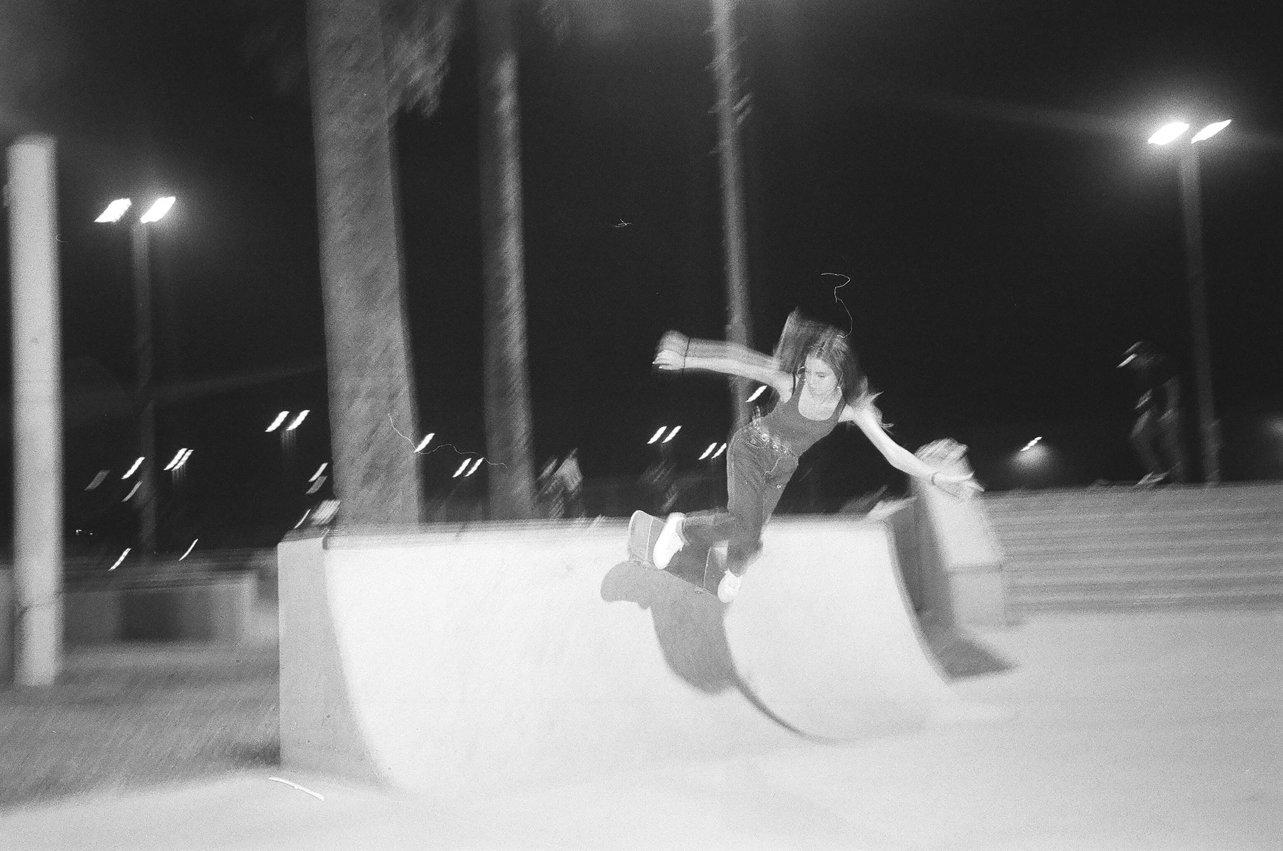 A person falling backward during a night skate at an outdoor skating rink, with trees and streetlights in the background.