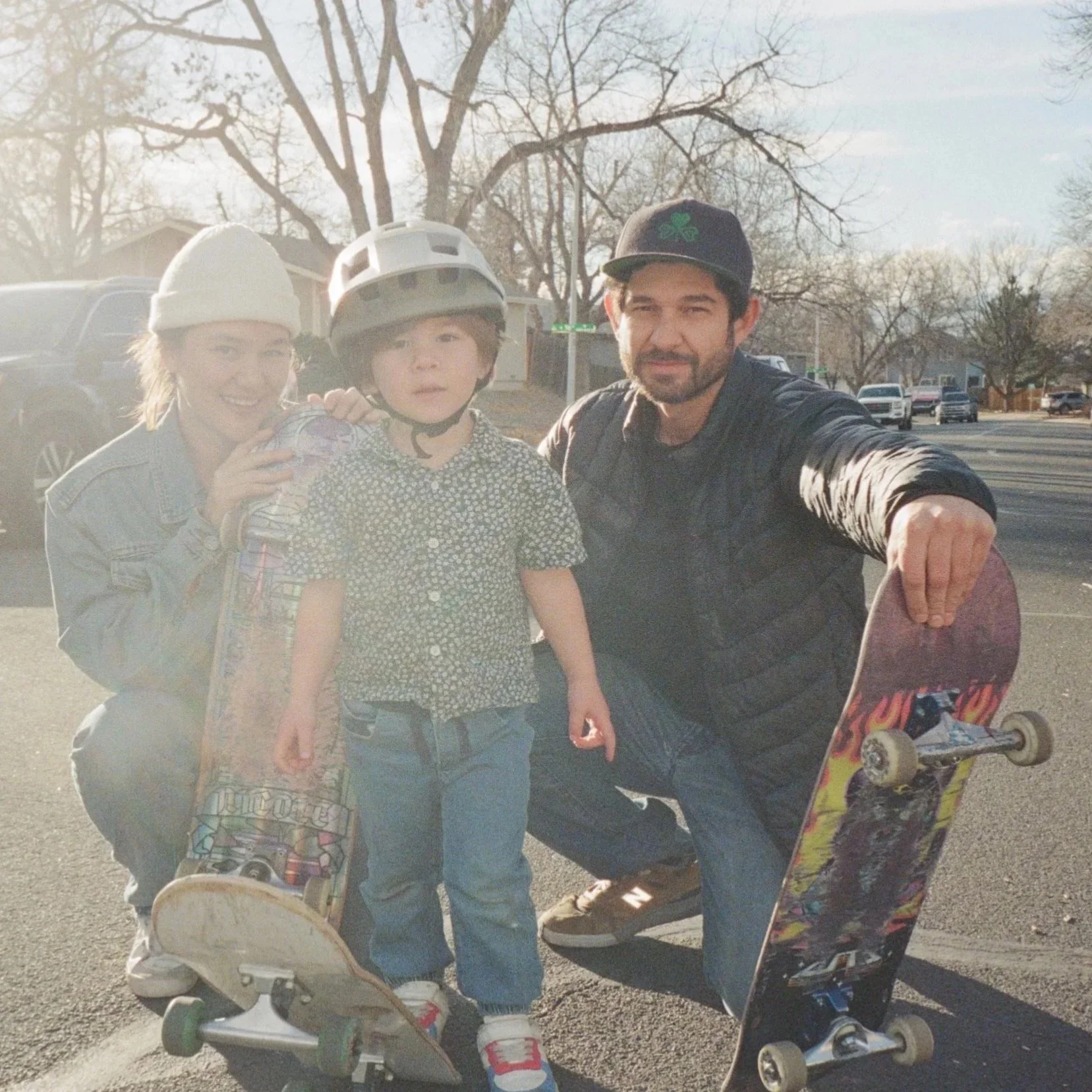 A family of three outdoors with skateboards, one child wearing a helmet, the parents kneeling next to their child, in a neighborhood street with parked cars and leafless trees.
