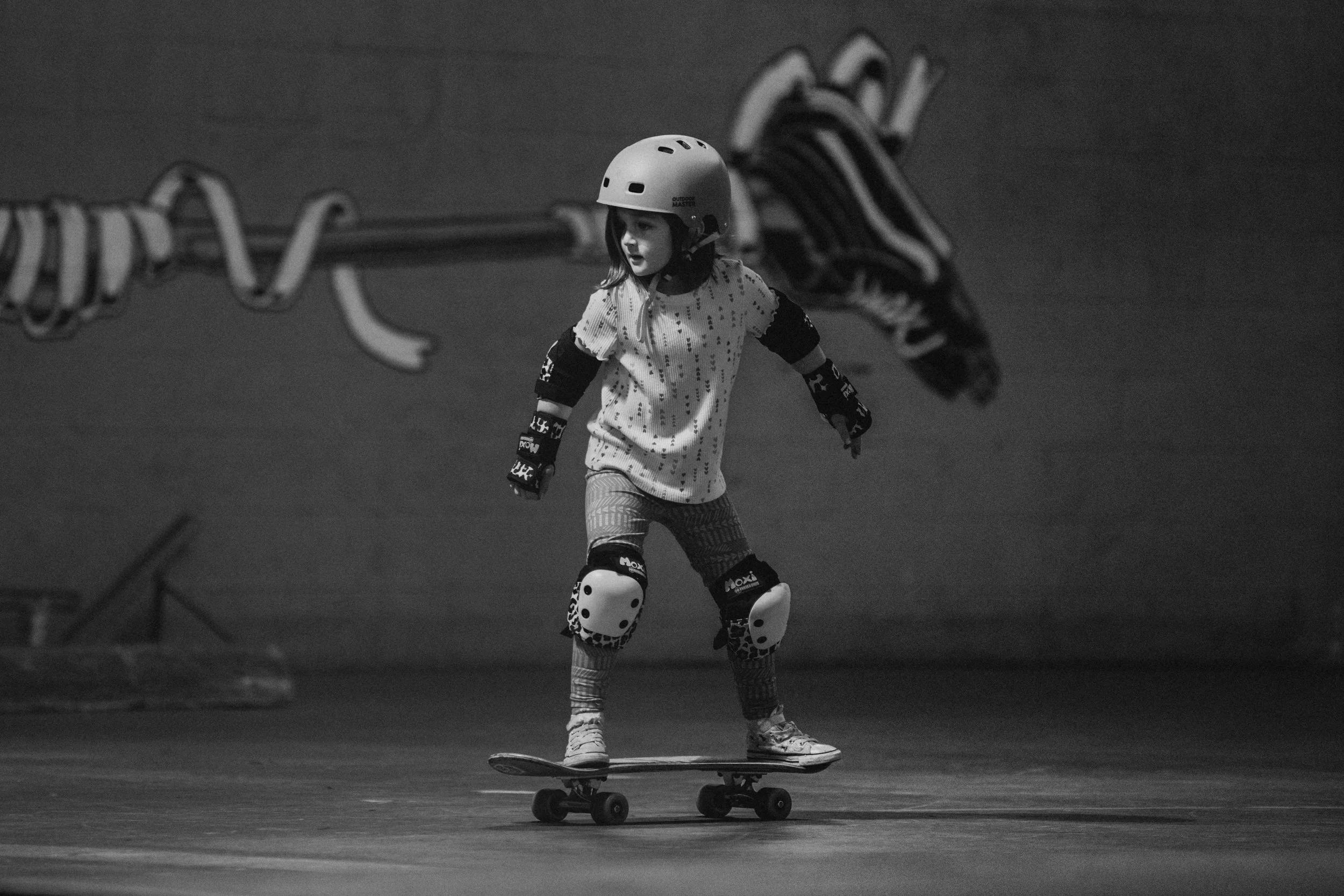 A young girl wearing a helmet and protective gear skateboarding indoors with a chain-link fence in the background.