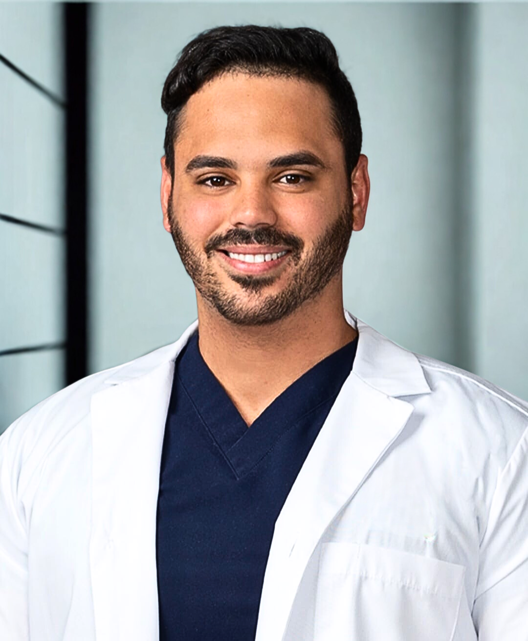 A smiling man with short dark hair, a beard, and light skin, wearing a white lab coat over navy blue scrubs, standing in a modern clinical setting.