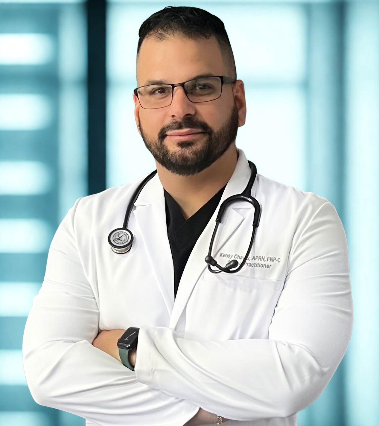 A male healthcare professional wearing glasses and a white lab coat with a stethoscope around his neck, standing with arms crossed in a medical facility.