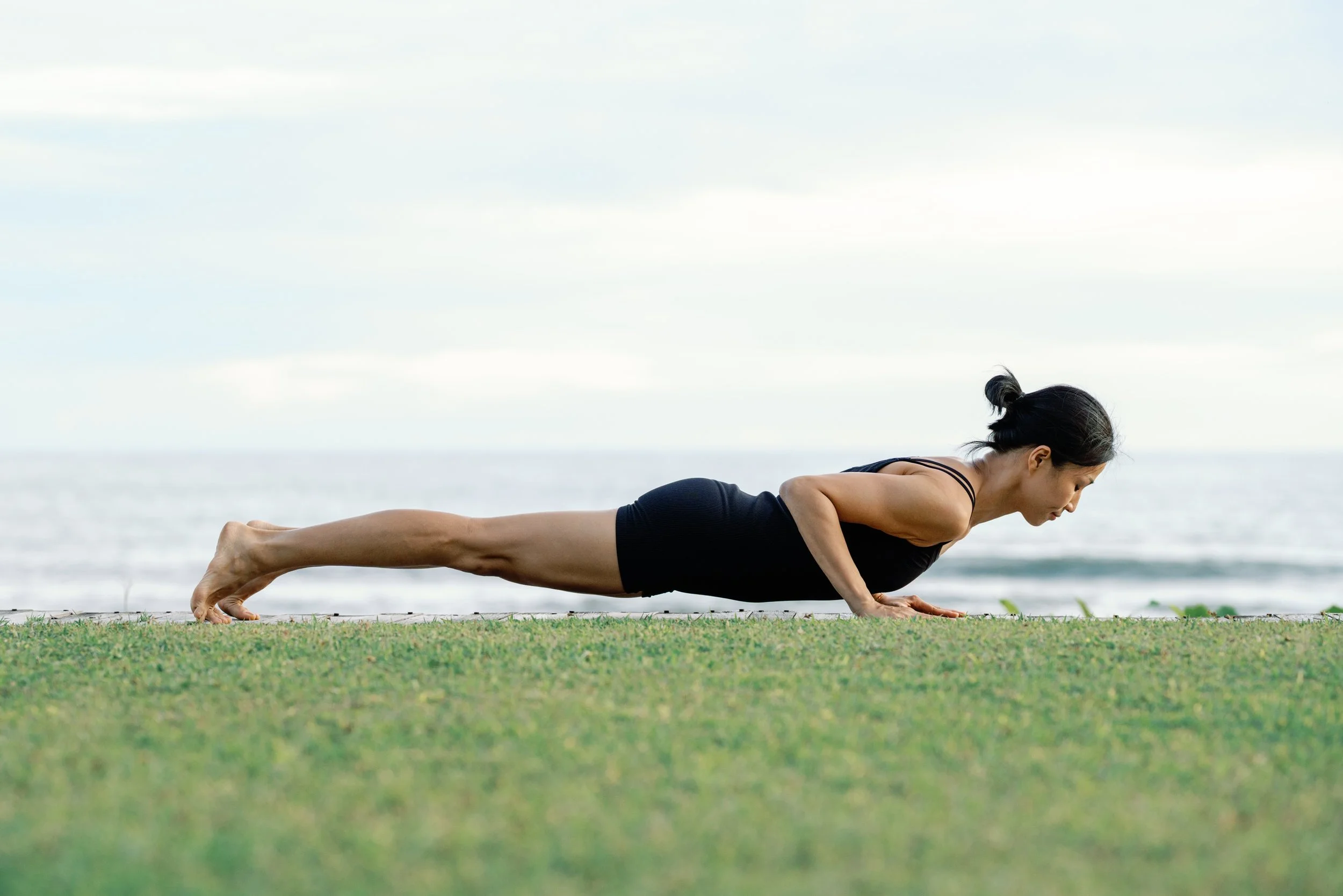 A woman practicing yoga outdoors near the ocean, holding a plank position on a grassy area.