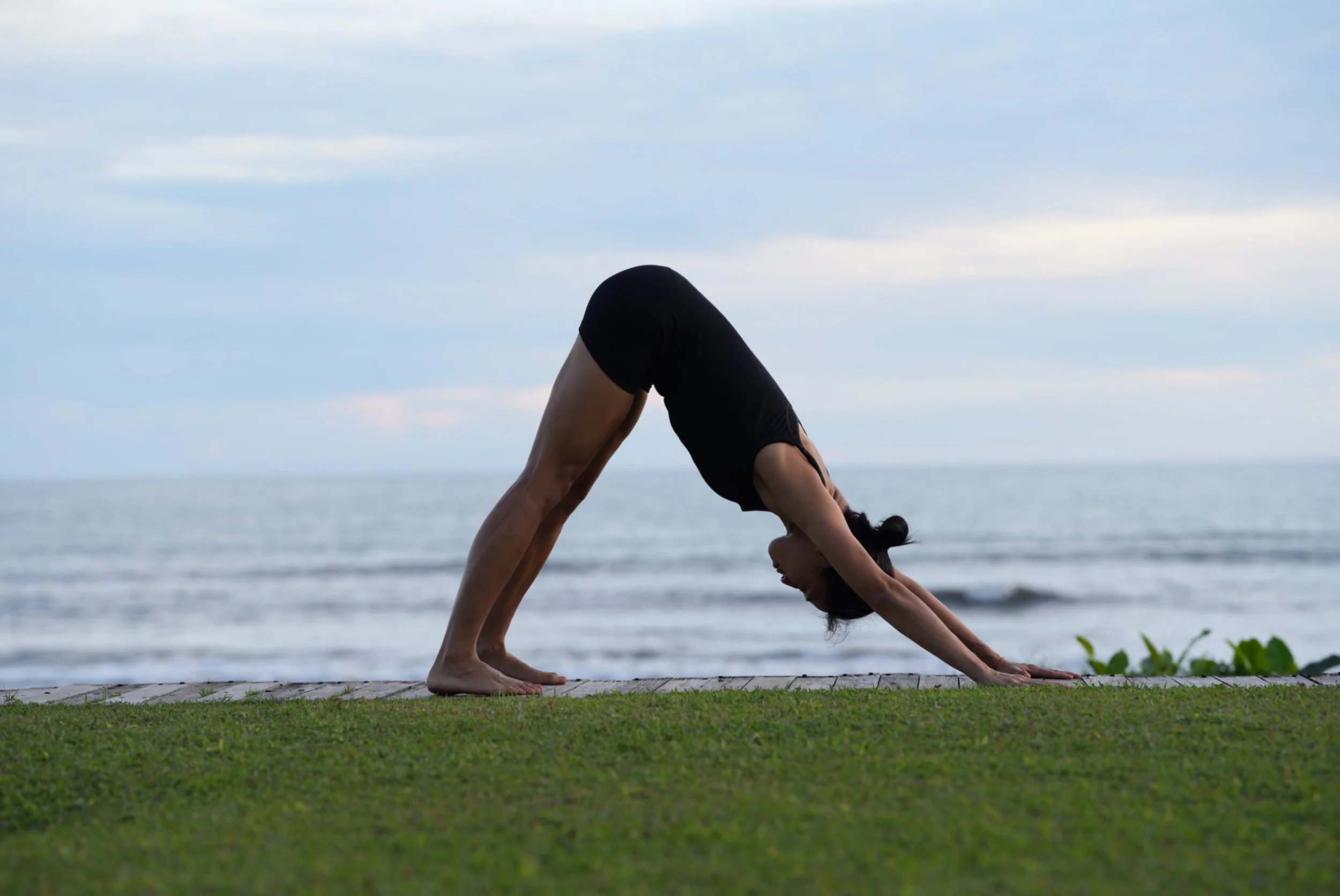 A woman doing downward dog yoga pose on grass near the ocean, with a cloudy sky above.