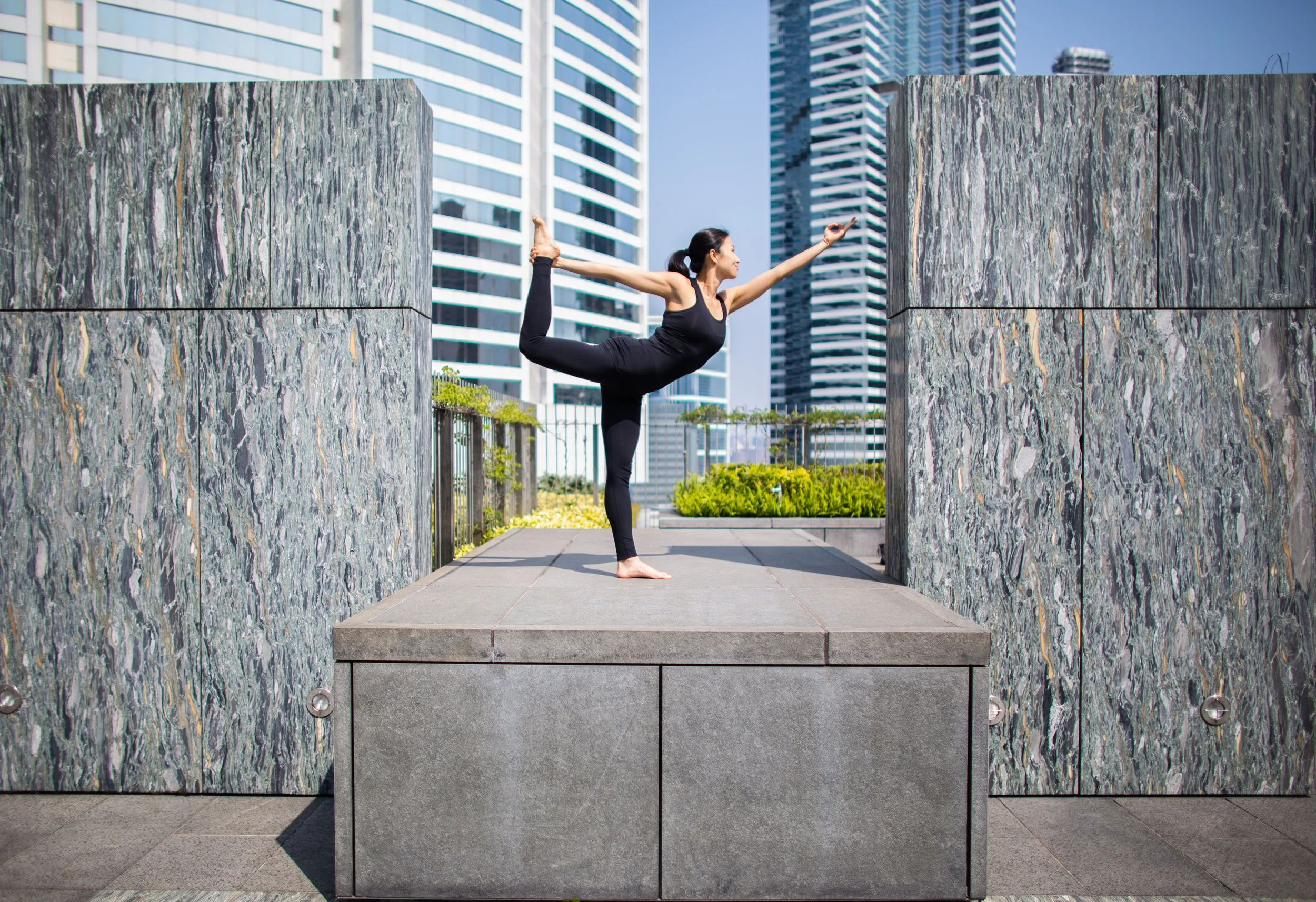 Woman in black athletic wear performing a yoga pose outdoors on a concrete platform with tall modern buildings in the background.