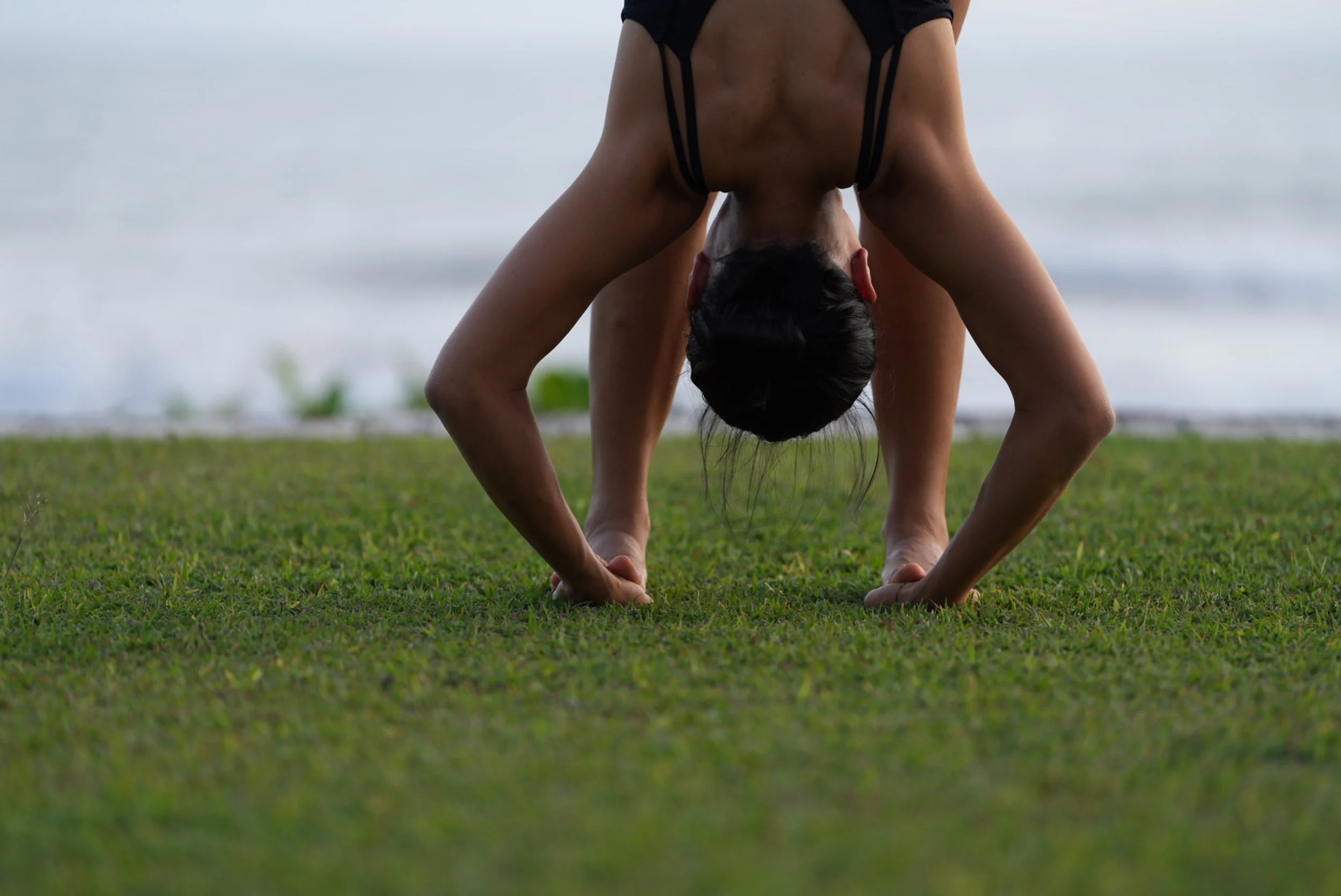 A person practicing yoga on grass near the beach, in a downward dog pose with hands and feet on the ground, head hanging downward.