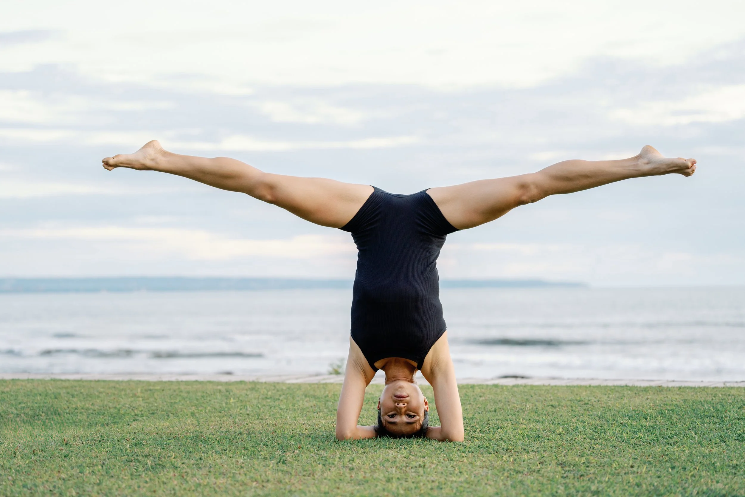 A child performing a handstand on grass near the ocean, with a cloudy sky overhead.