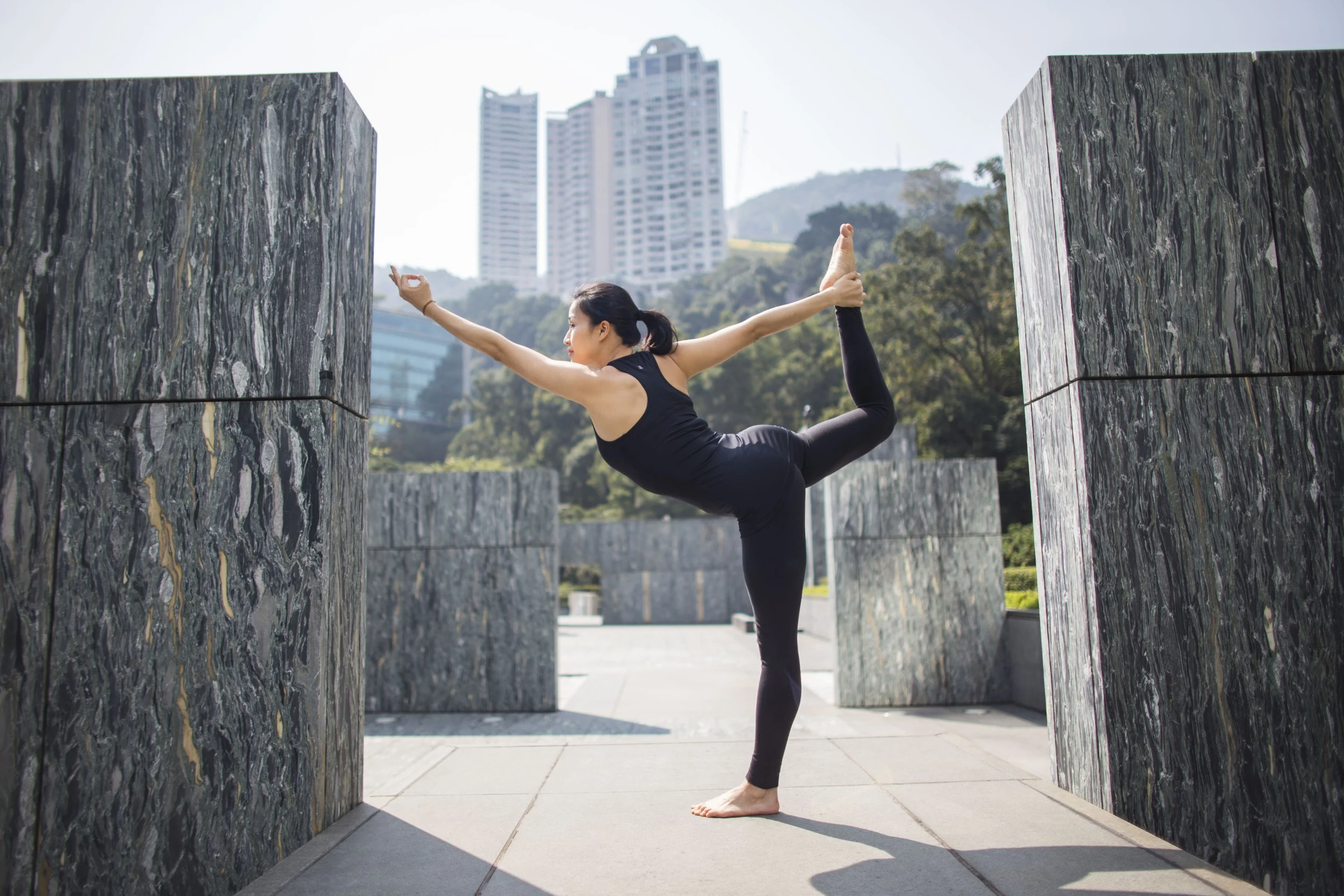 A woman practicing yoga outdoors, performing a balancing pose on one leg with arms extended, between large marble-like stone structures, with tall buildings and greenery in the background.