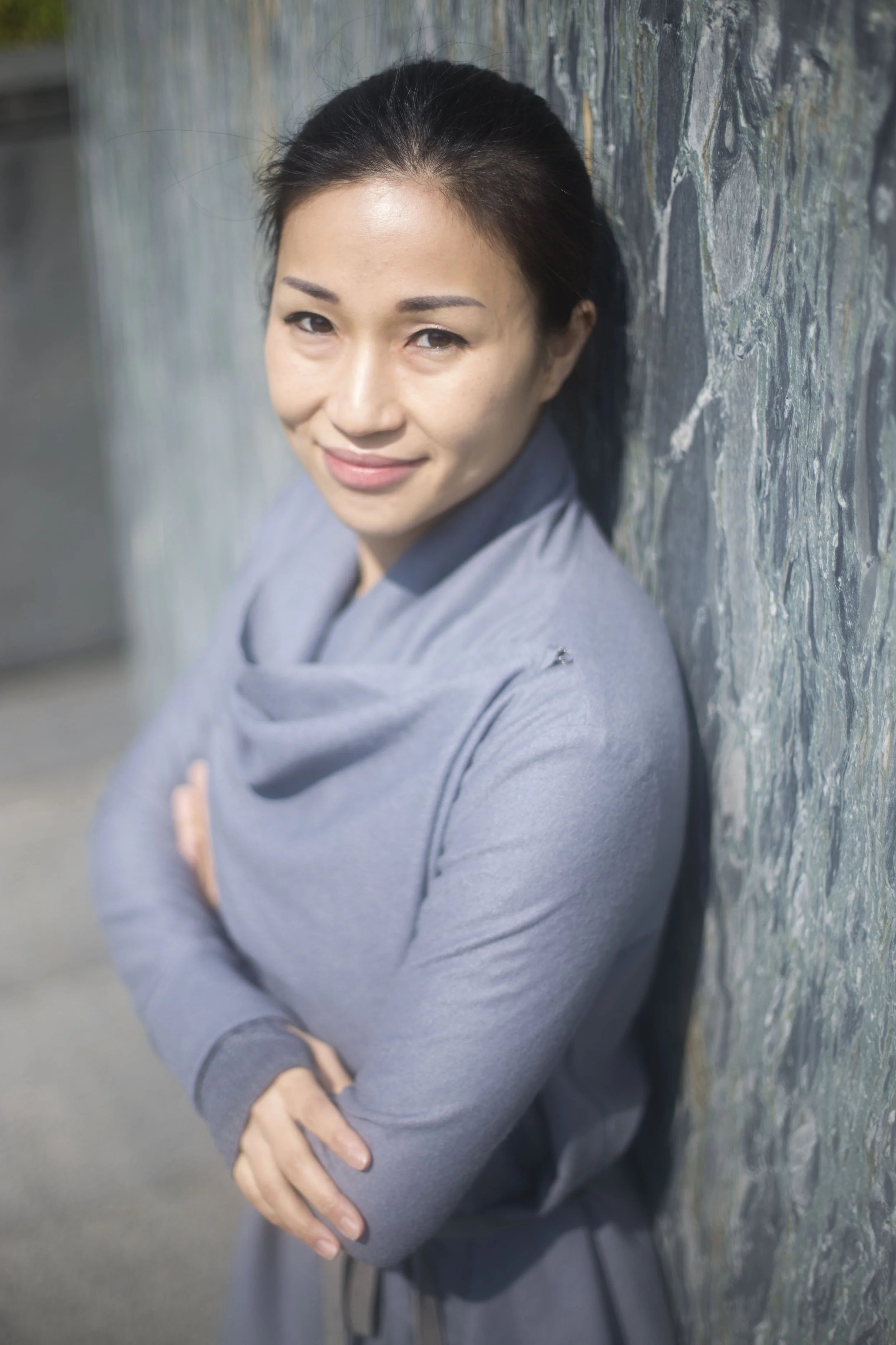 A woman with dark hair pulled back, smiling with crossed arms, leaning against a textured stone wall in daylight.