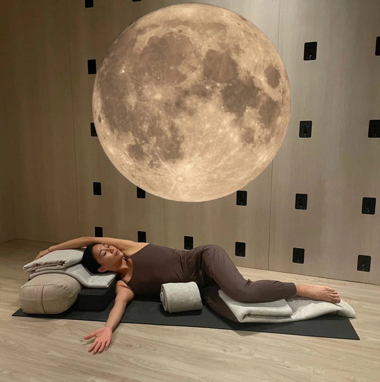 A woman lying on a mattress with pillows and blankets, using a moon-shaped object as a pillow, in a room with a large moon wall decoration overhead.