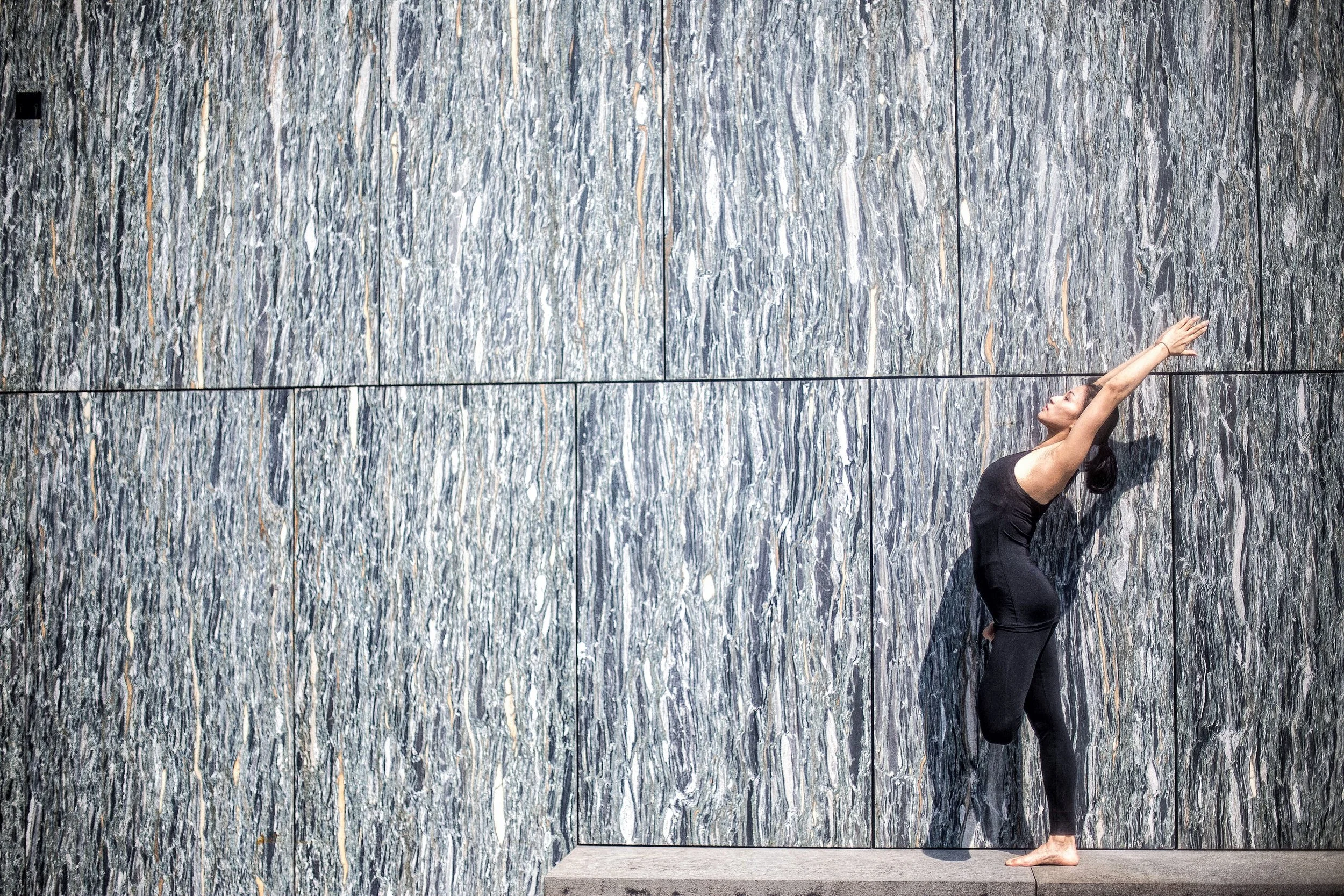 A woman in a black outfit practicing yoga or stretching against a large gray stone wall with a textured surface.