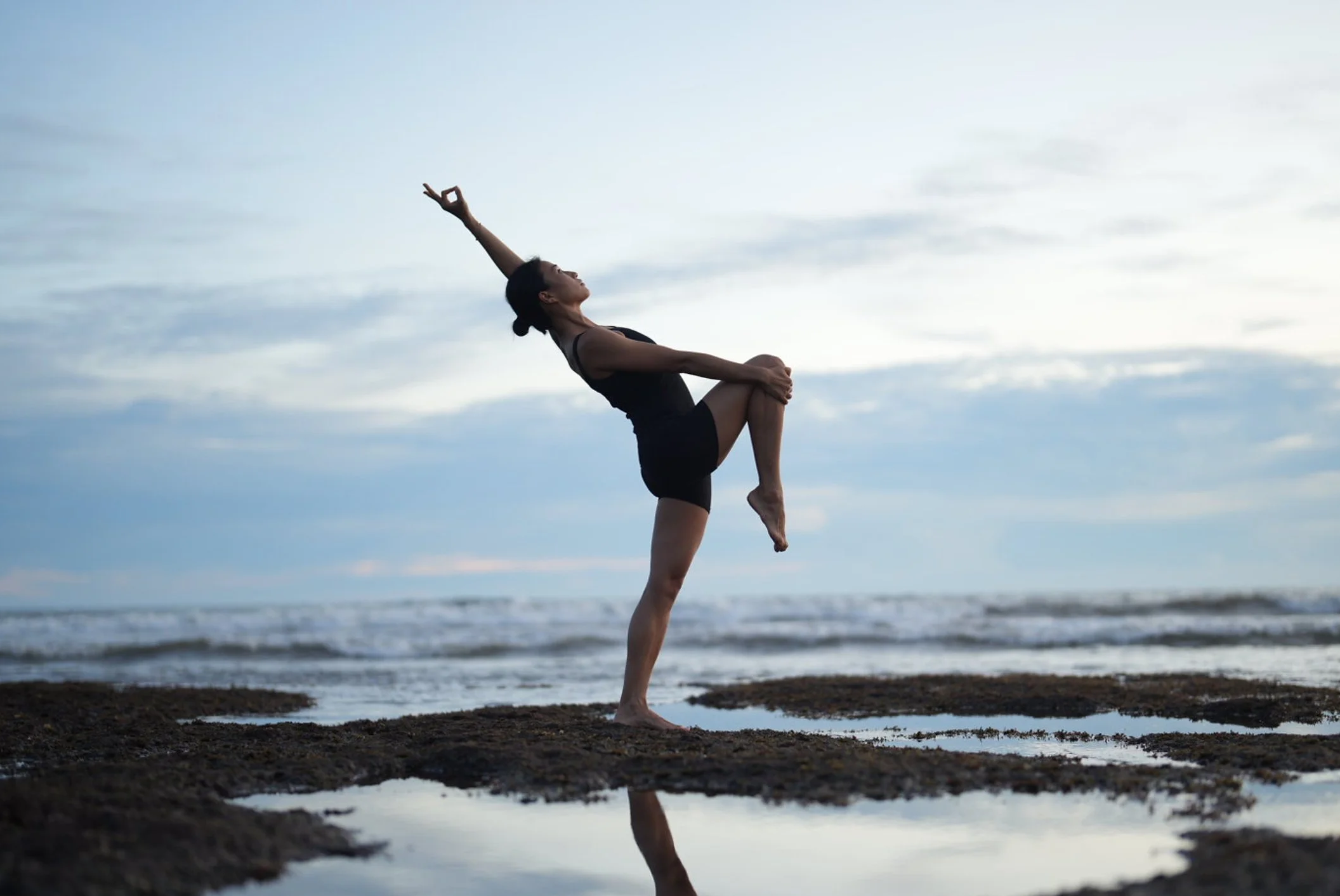 A woman practicing yoga on the beach during sunset, standing on one leg with her arms raised and mirroring her reflection in a puddle of water.