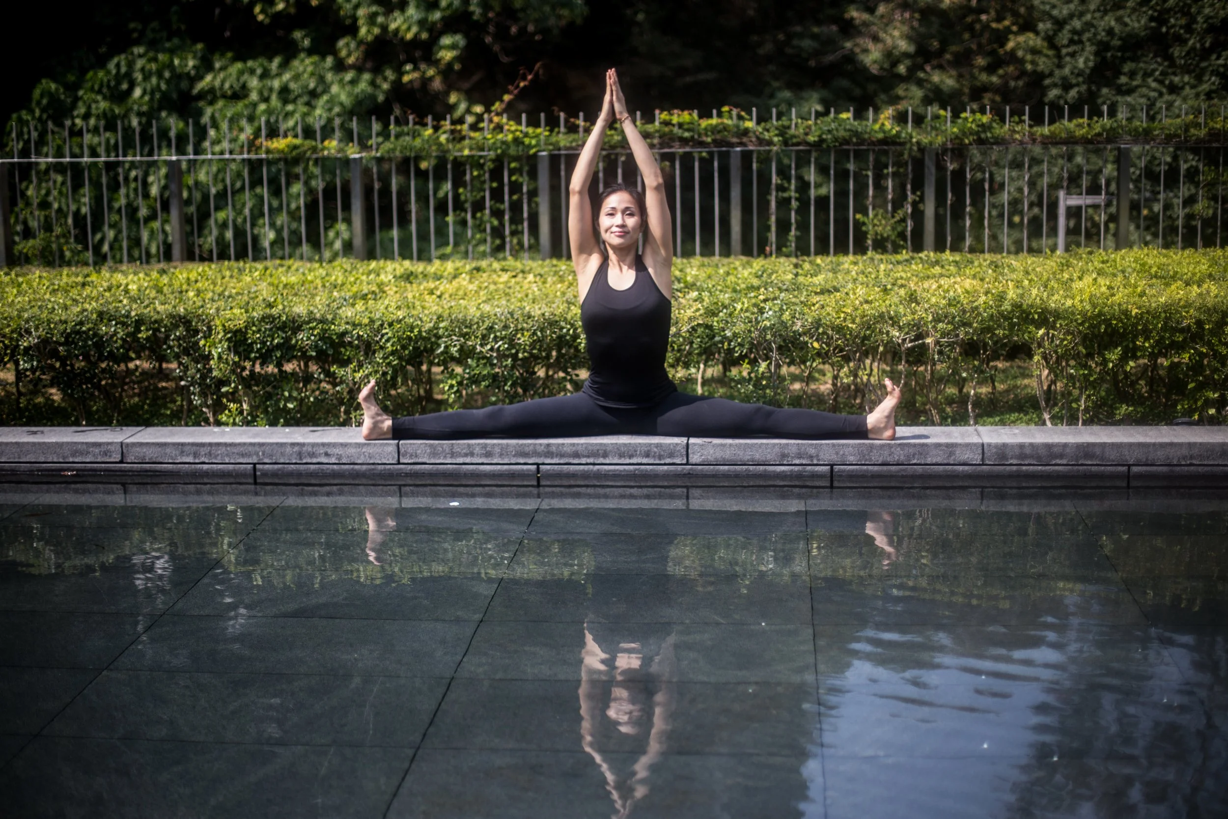 Woman practicing yoga by a pool, sitting in a split position with arms raised above her head outdoors.