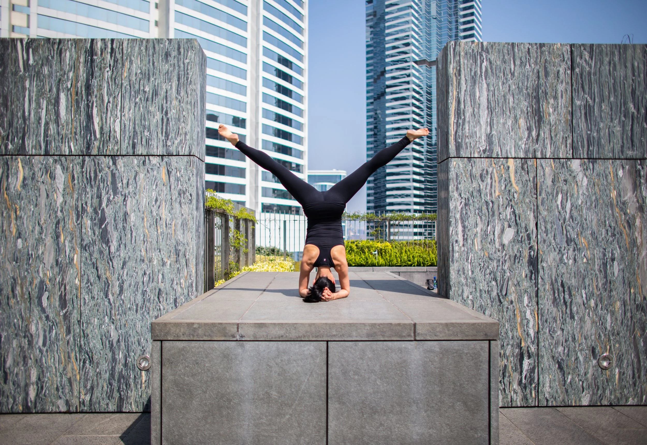 A woman performing a headstand yoga pose outdoors on a rectangular stone platform, with her legs spread in a V shape. She is dressed in black athletic wear, with modern skyscrapers in the background.