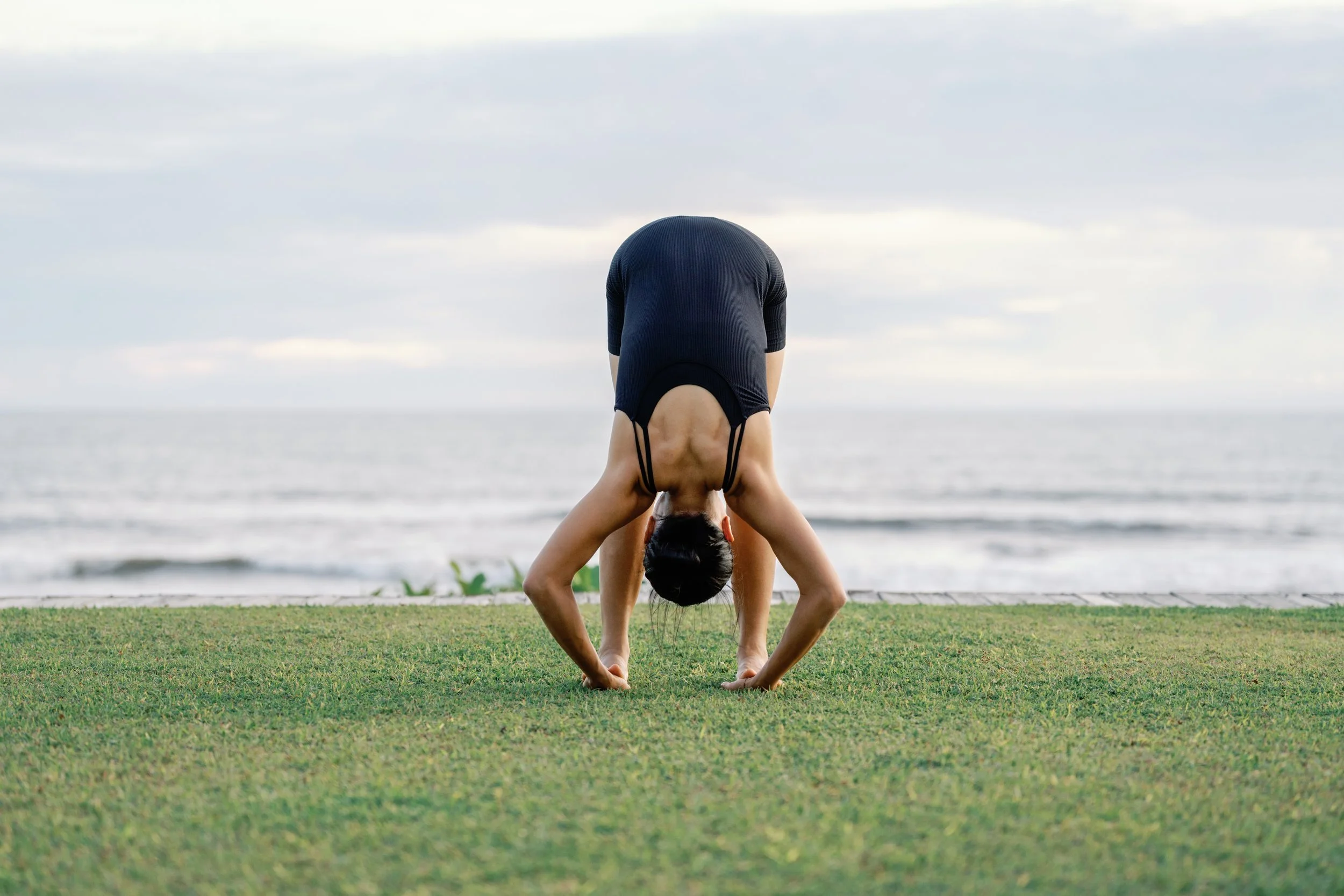 A person performing a downward dog yoga pose on green grass near the ocean, with cloudy sky above.