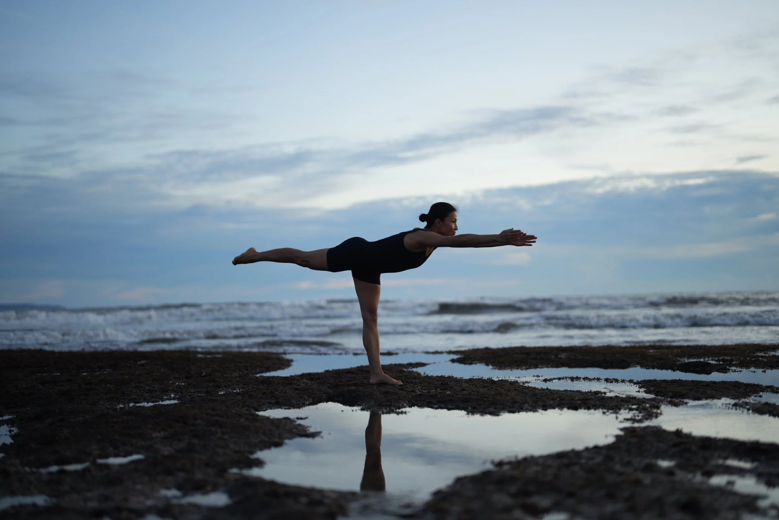 A woman practicing a yoga pose on a beach during sunset or dusk, balancing on one leg with the other leg extended backward and arms stretched forward.