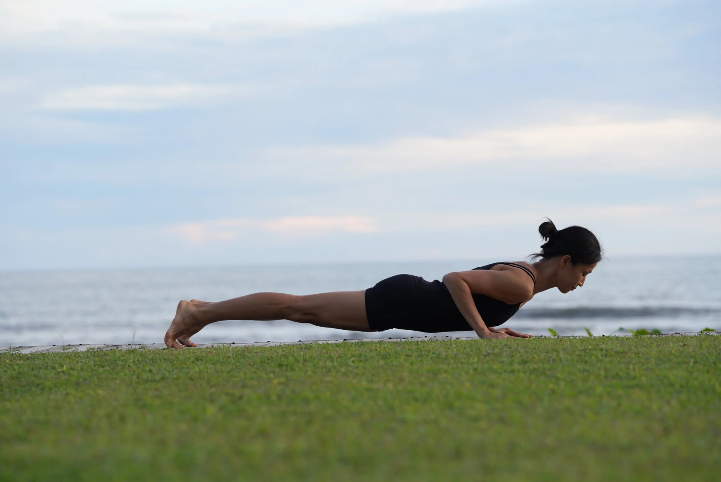 A woman practicing yoga outdoors near the ocean, performing a plank pose on a grassy surface.