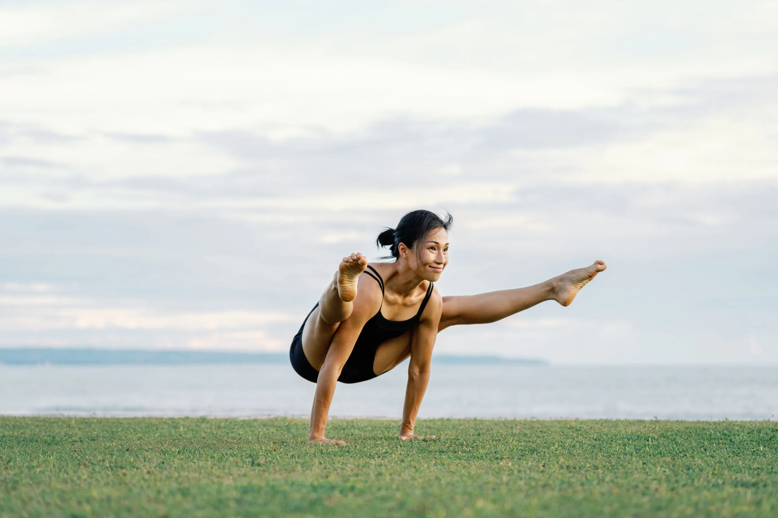 A woman practicing yoga outdoors, balancing on her hands with her legs extended in a split position against a backdrop of a cloudy sky and a body of water.